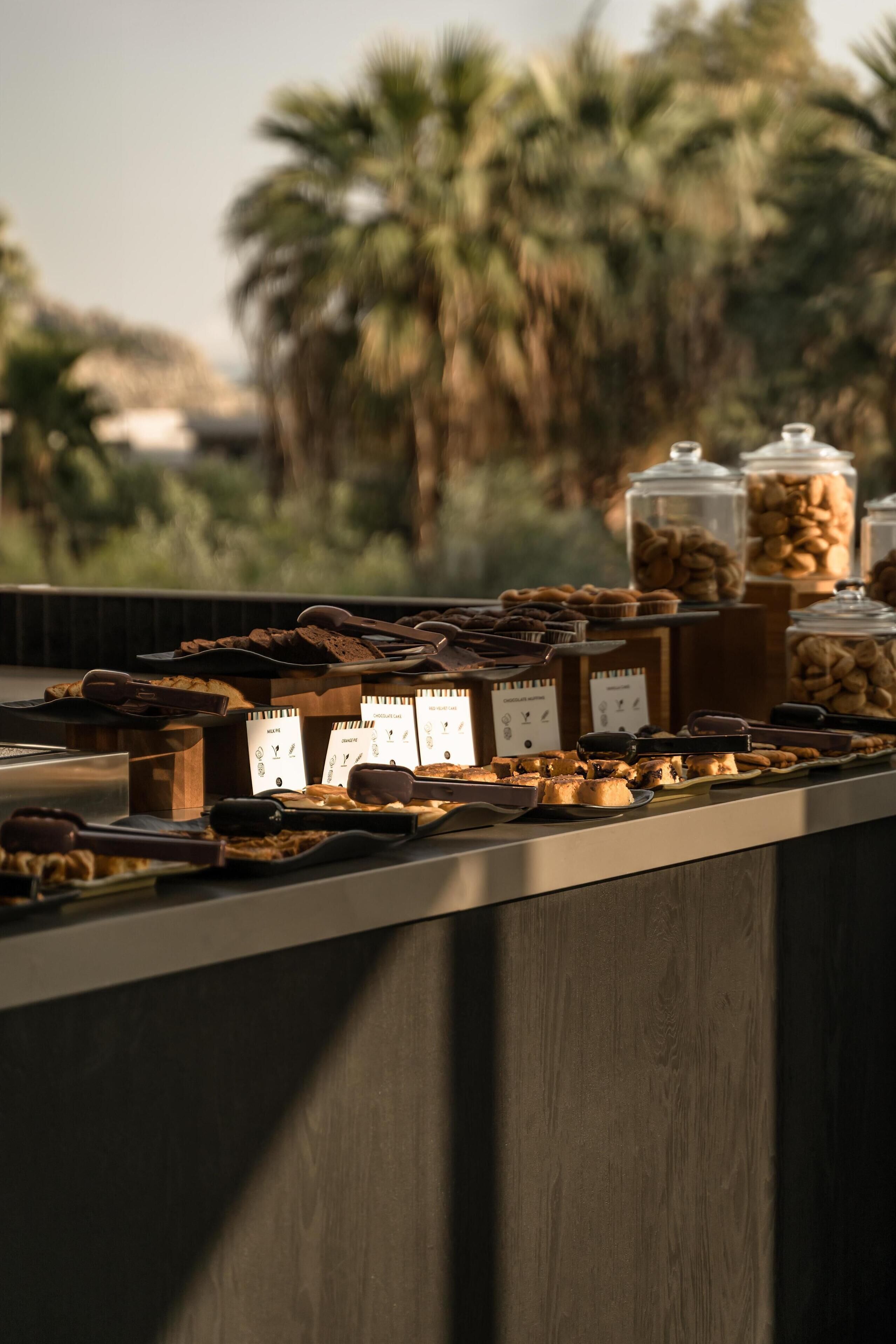 Un bufé con diversos productos horneados y pasteles, con vistas a palmeras al fondo.