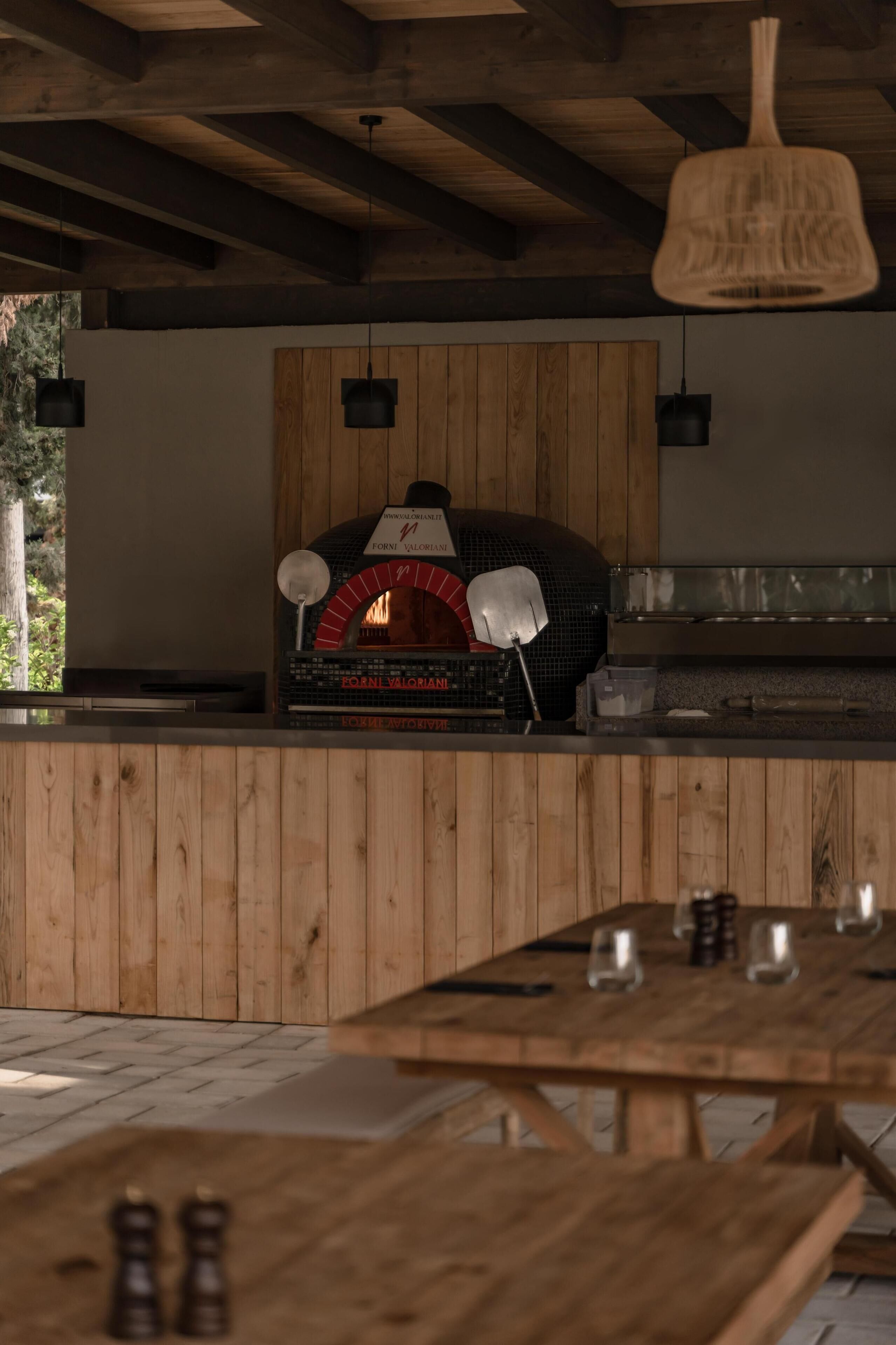 Een houten interieur met een bakoven, houten paneelwanden, hangende lampen en een tafel met glazen.