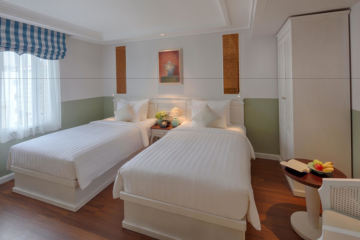 A hotel room with two beds, a nightstand with a lamp, a small table with fruits and a book, and a wardrobe.