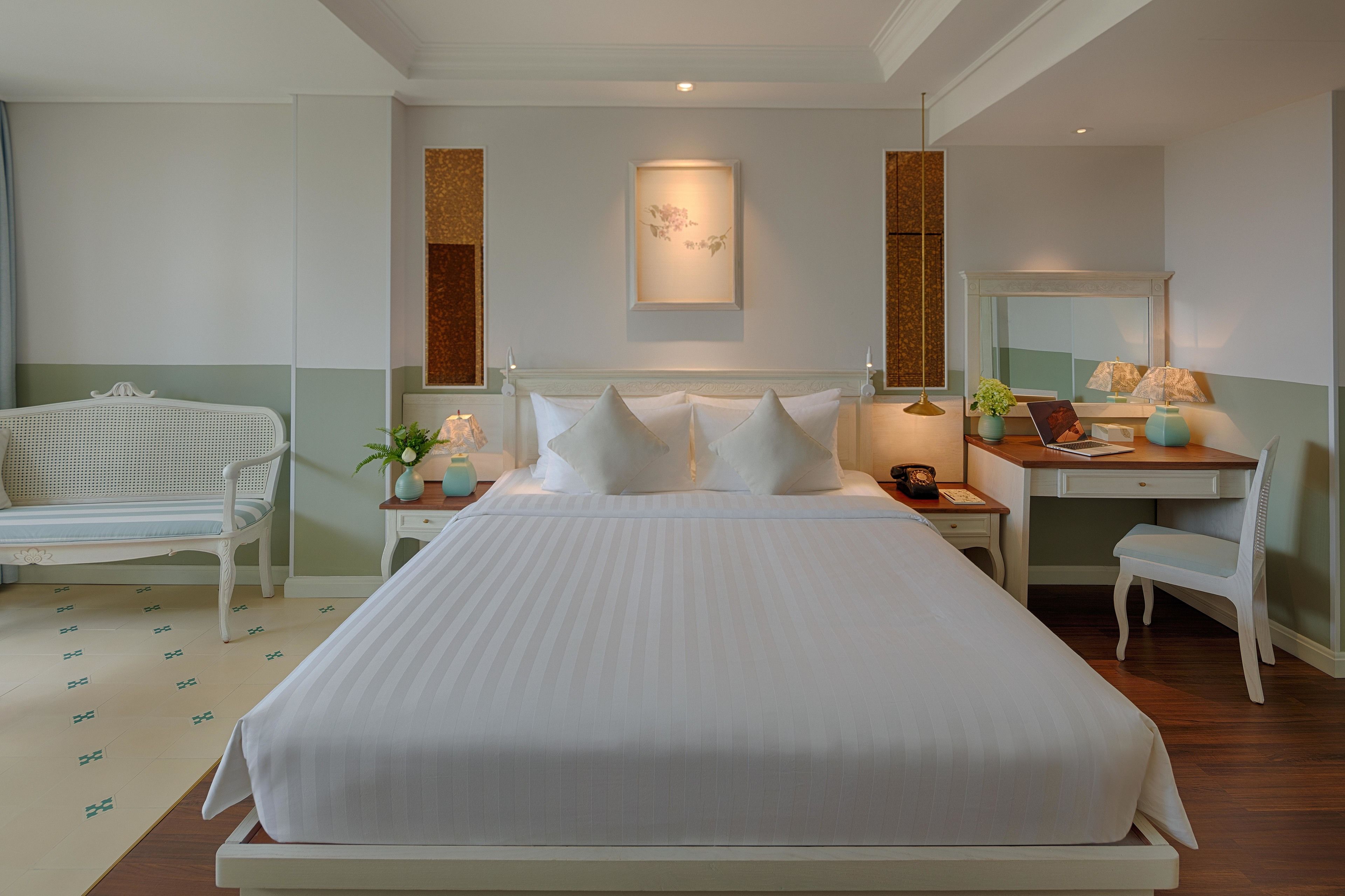 A neatly arranged hotel room with a large bed, a desk with a chair, a small table with a plant, and a mirror.