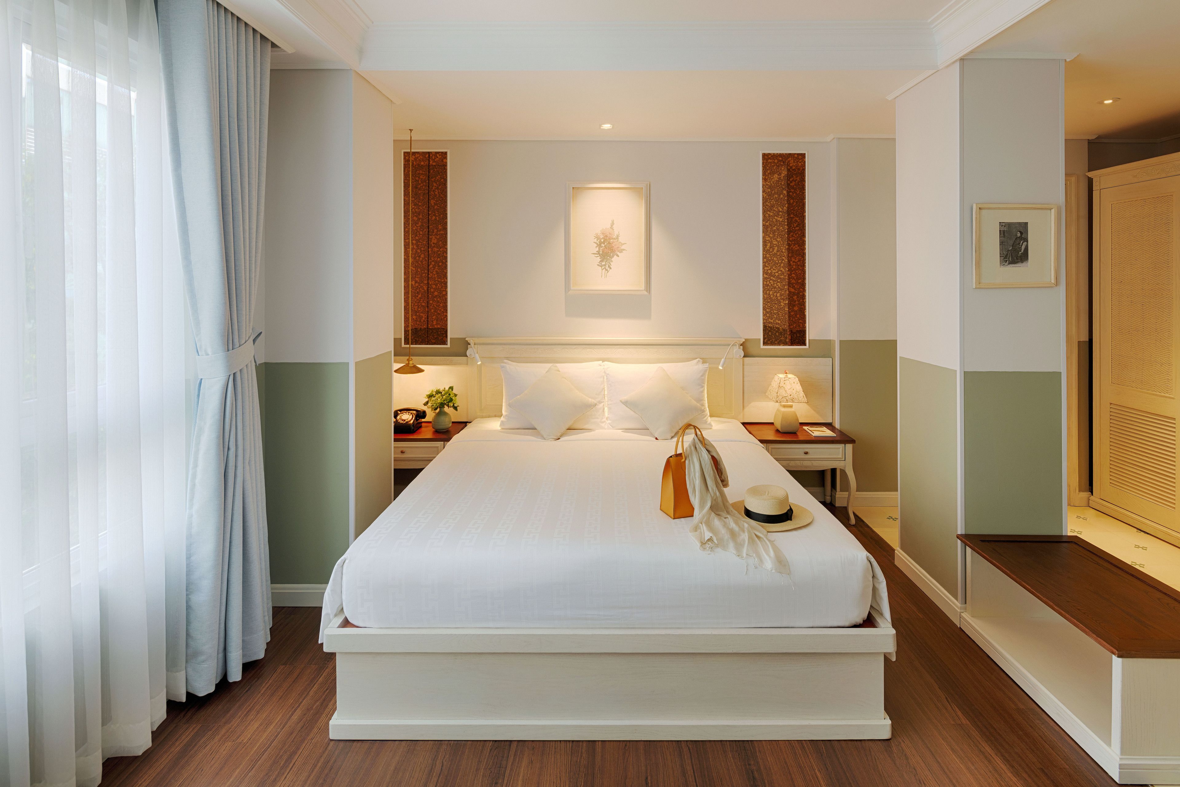Una cama grande con ropa de cama blanca, dos mesitas de noche con lámparas, un cuadro en la pared y un banco de madera junto a la cama.