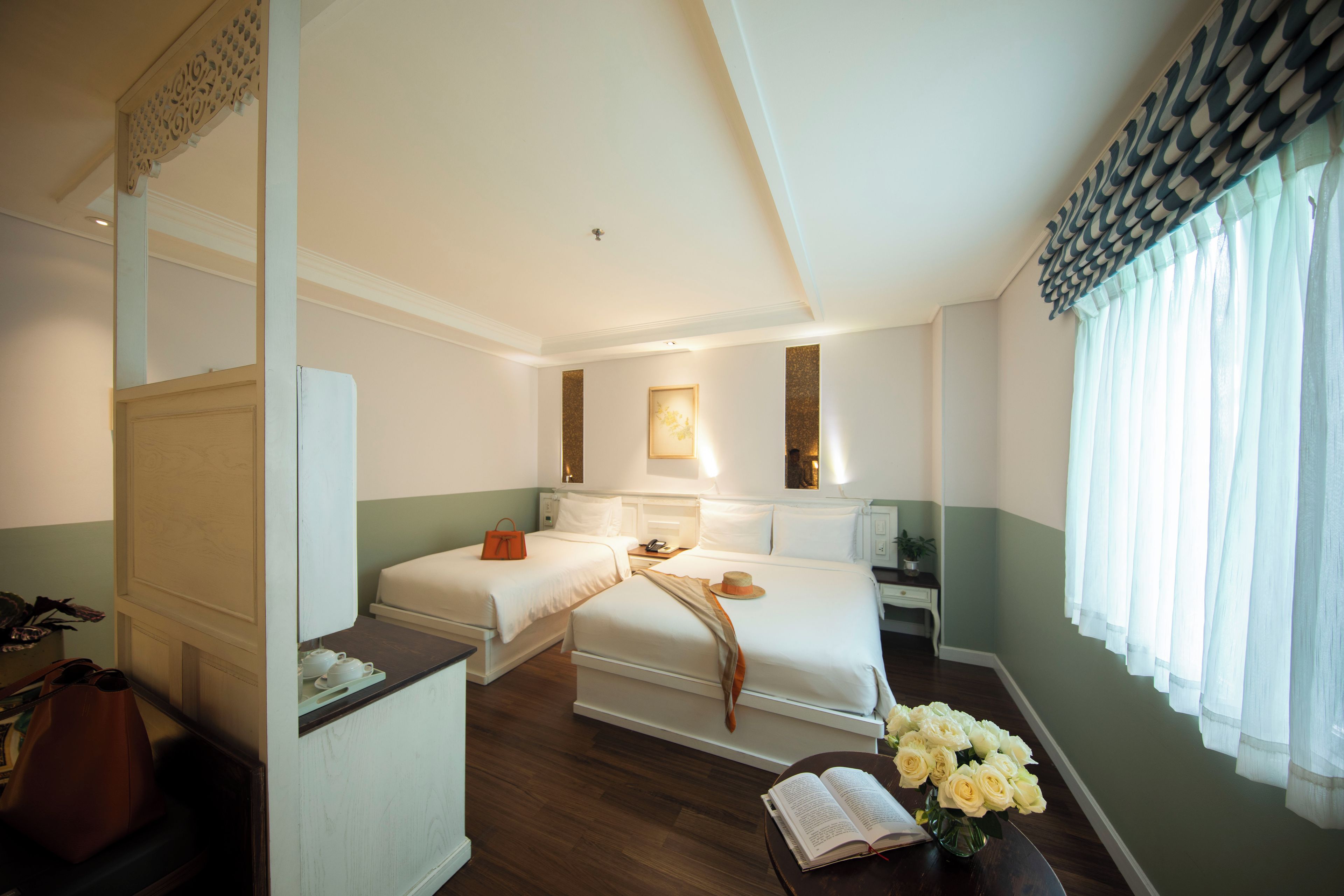 Habitación de hotel con dos camas, un escritorio, una silla, una mesita con un jarrón de flores y una ventana con cortinas.