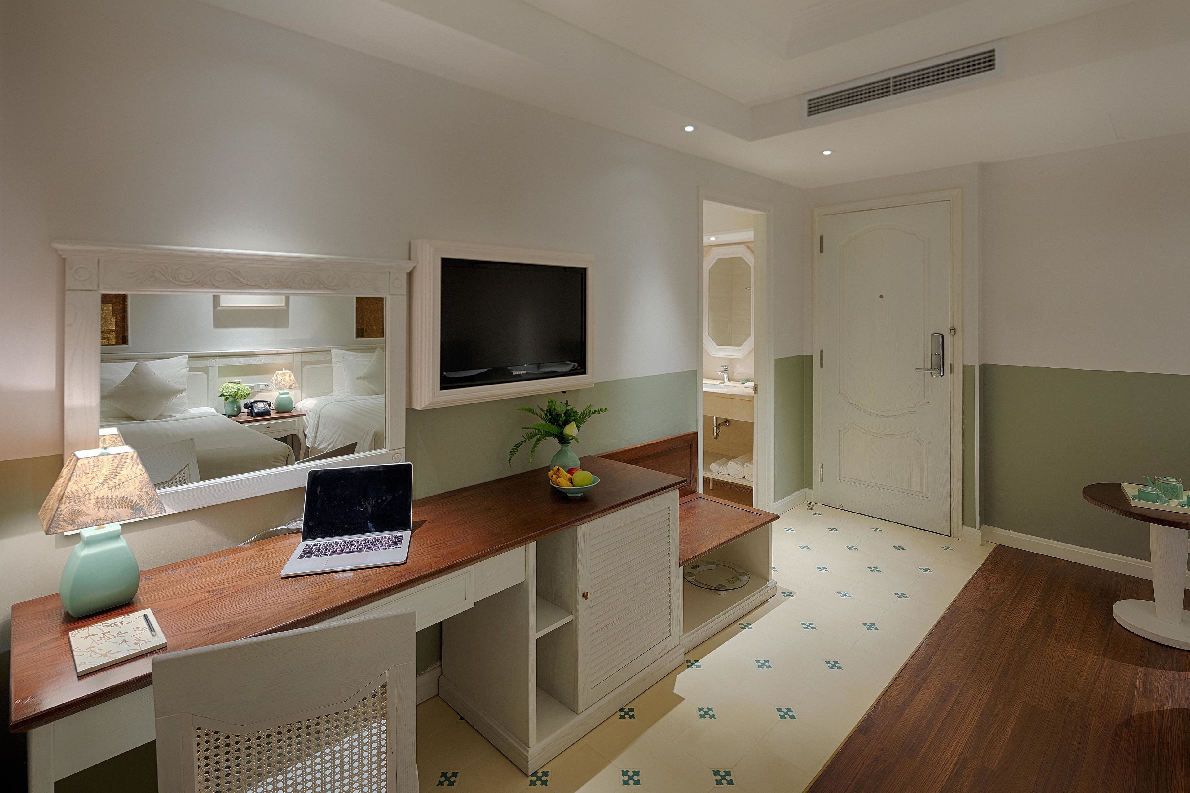 Une chambre d’hôtel avec un bureau en bois, un ordinateur portable, un miroir, une télévision et une petite table sur laquelle se trouvent un vase et des fruits.