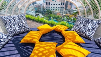 A sunlit outdoor seating area with patterned cushions, a view of a cityscape, and a pool in the background.