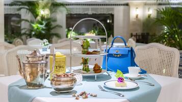 Ein Tisch ist für eine Teezeremonie gedeckt, mit einem Etageren-Kuchenständer, einem Teekessel, Zuckerdose, Milchkännchen, einer blauen Handtasche, einem Teller mit einem Kuchenstück und einer Tasse.