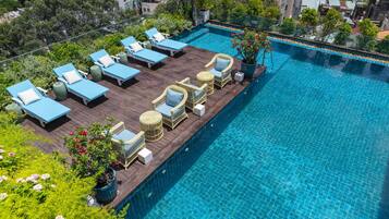 A rooftop swimming pool with lounge chairs and wicker furniture, surrounded by greenery and urban buildings.