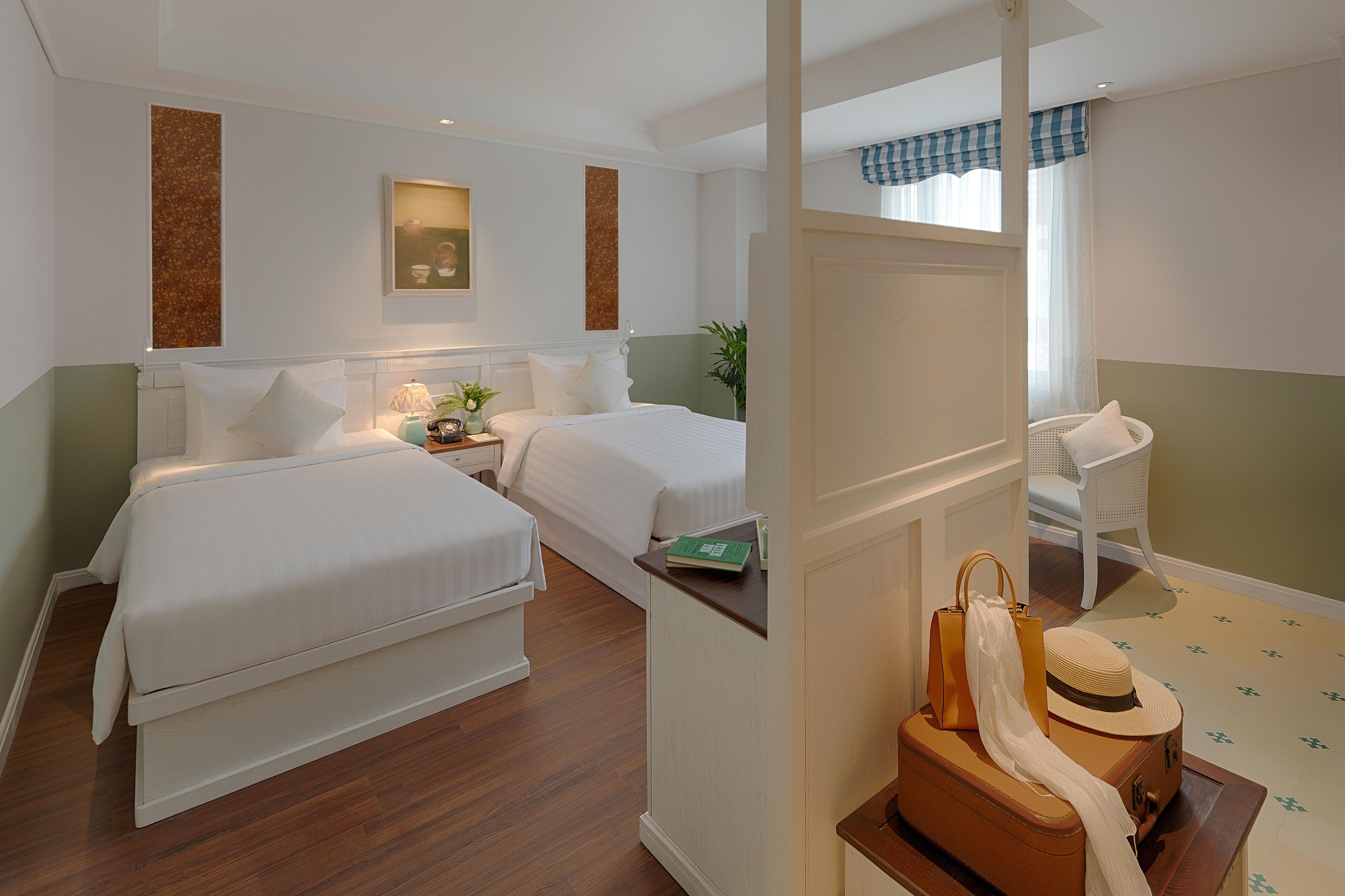 A hotel room with two beds, a small table with a book and a handbag, a chair, and a suitcase on the floor.