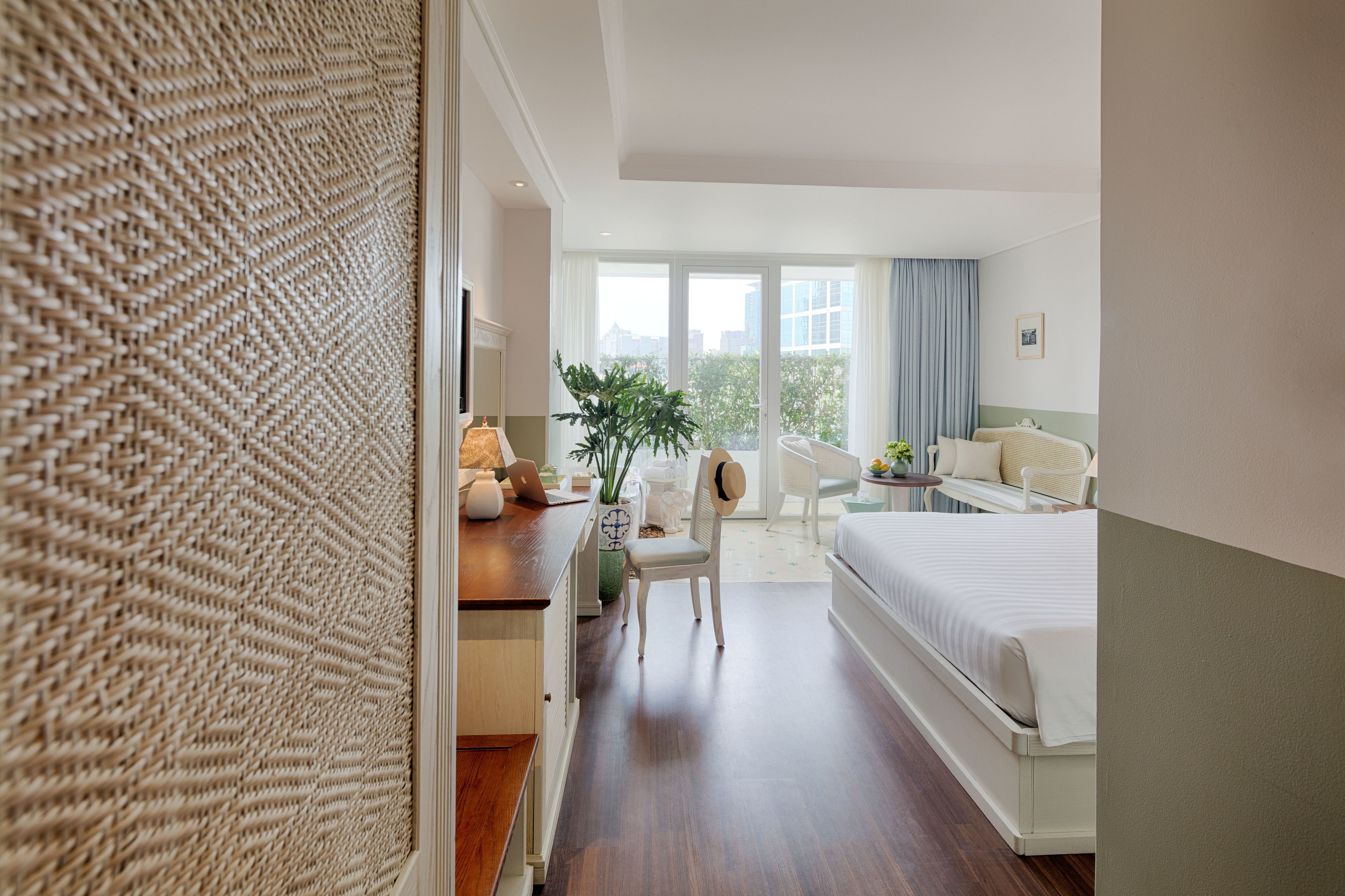 A hotel room with a bed, a desk with a laptop, a chair, a plant, and a view of the city through large windows.