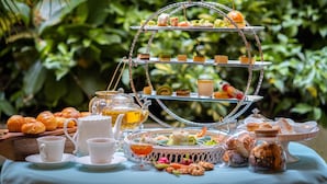 A three-tiered dessert stand with various pastries and a teapot on a table with a blue tablecloth, surrounded by greenery. - Au Lac Charner Hotel (Ho Chi Minh City)