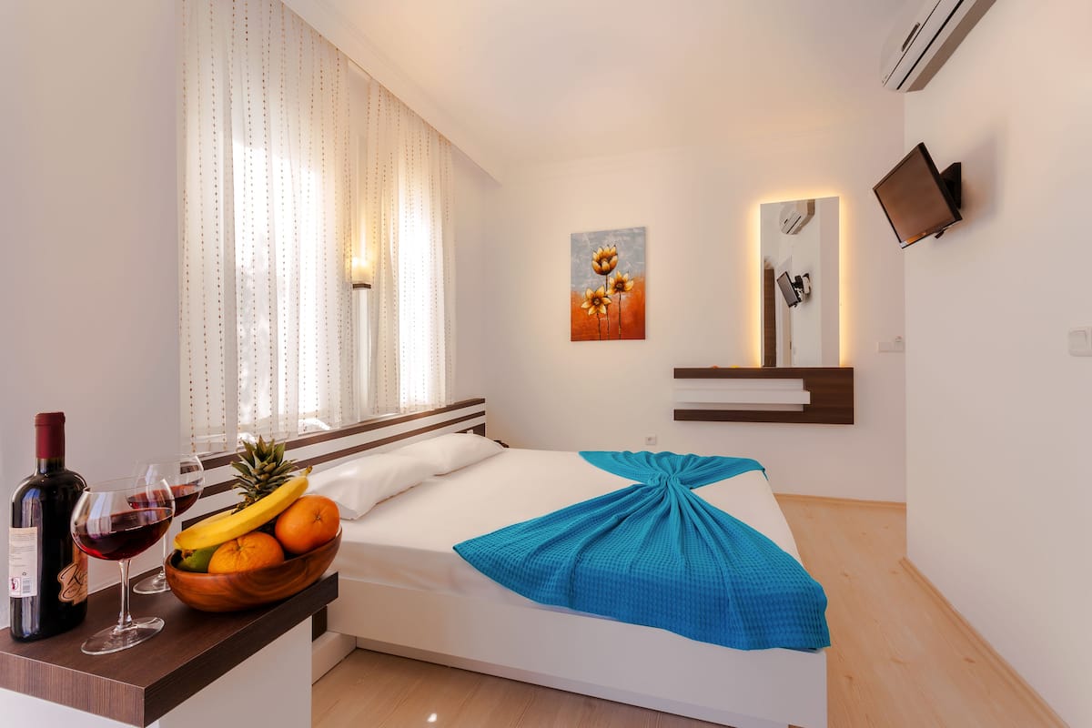 Habitación de hotel con cama, mesita de noche con una botella de vino, vasos y un frutero. Hay un cuadro en la pared y un televisor montado allí mismo.