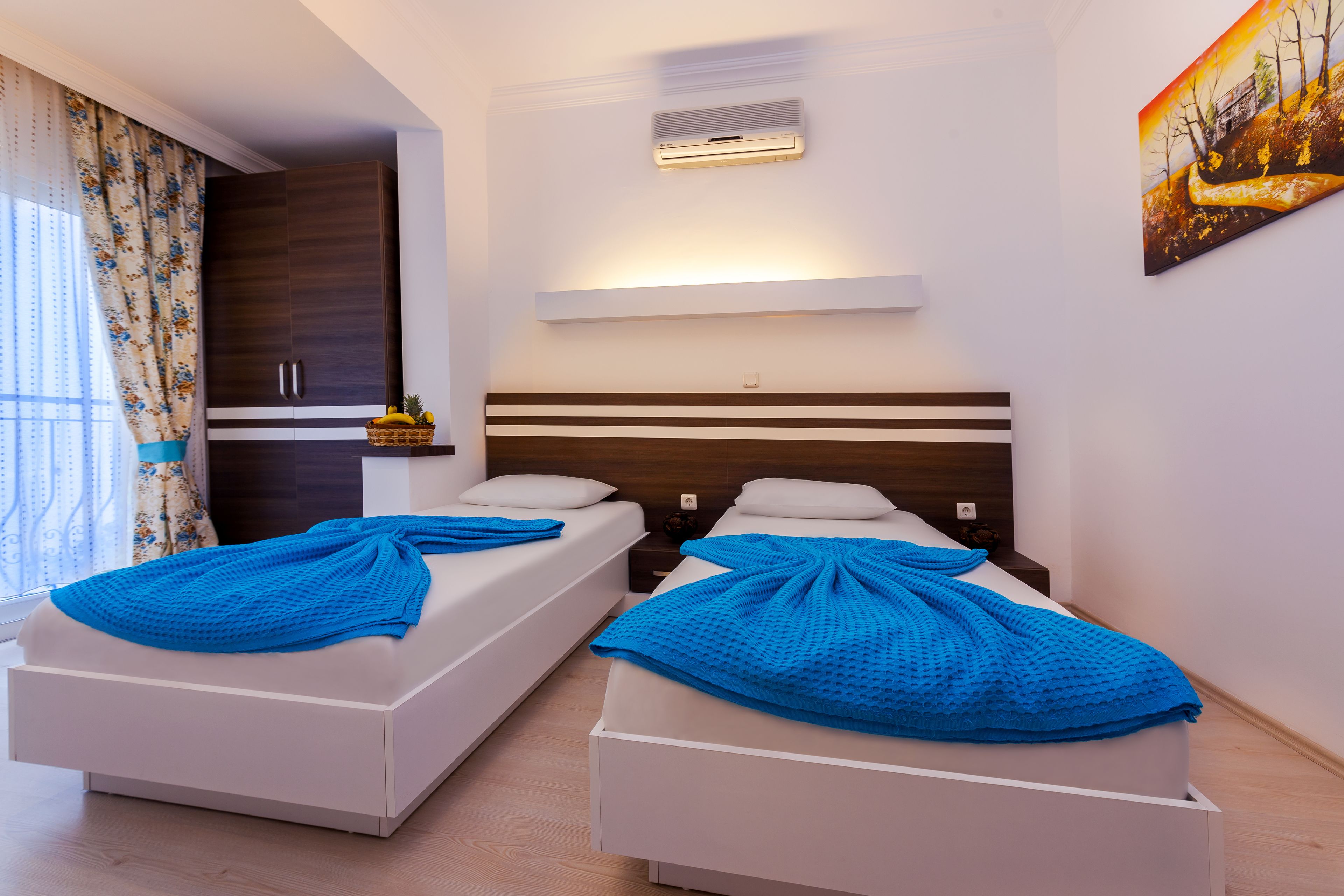 A hotel room with two beds, a large window with curtains, a painting on the wall, and a ceiling-mounted air conditioner.