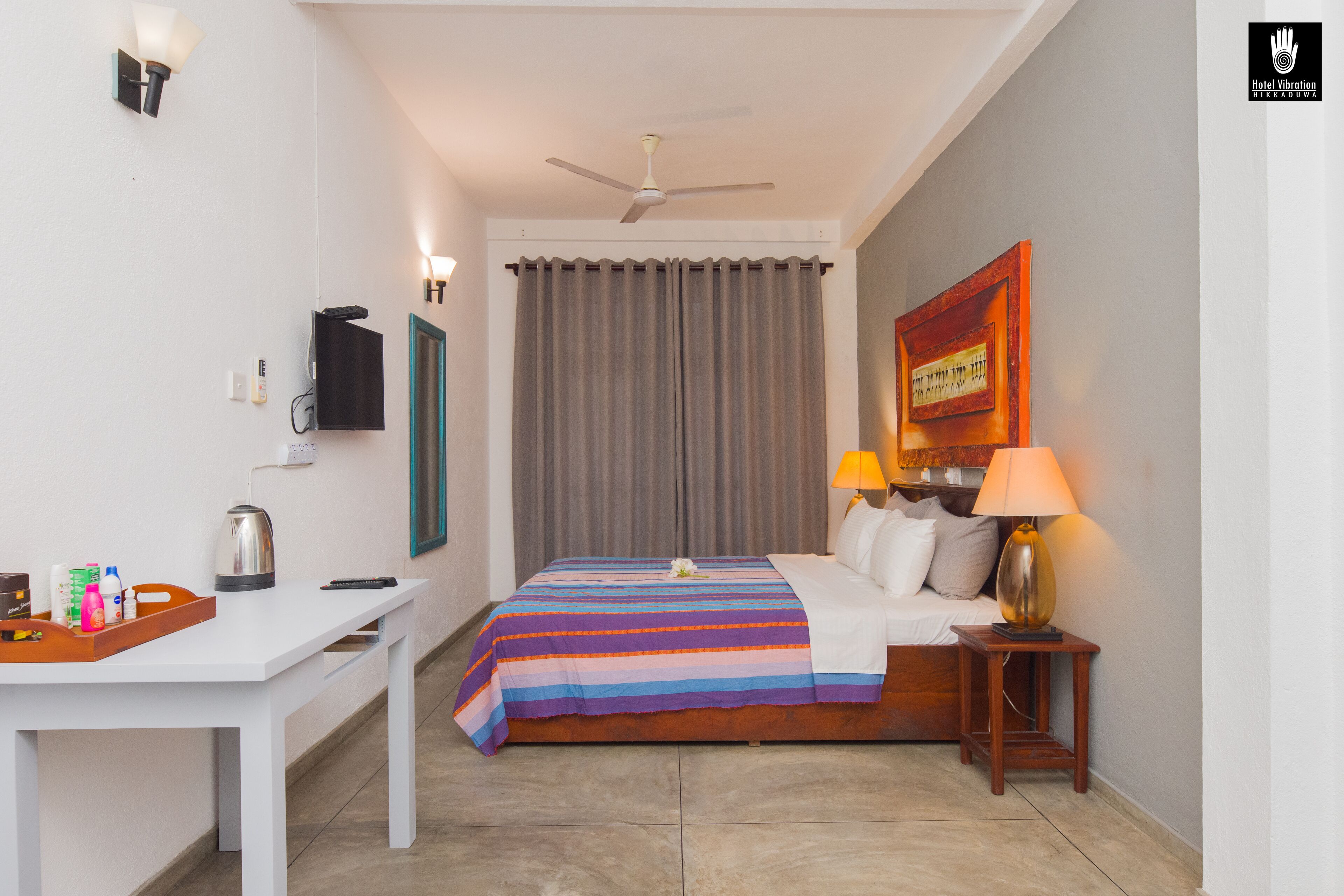 A hotel room with a bed, a desk, a chair, a nightstand, a lamp, a ceiling fan, and a curtain.