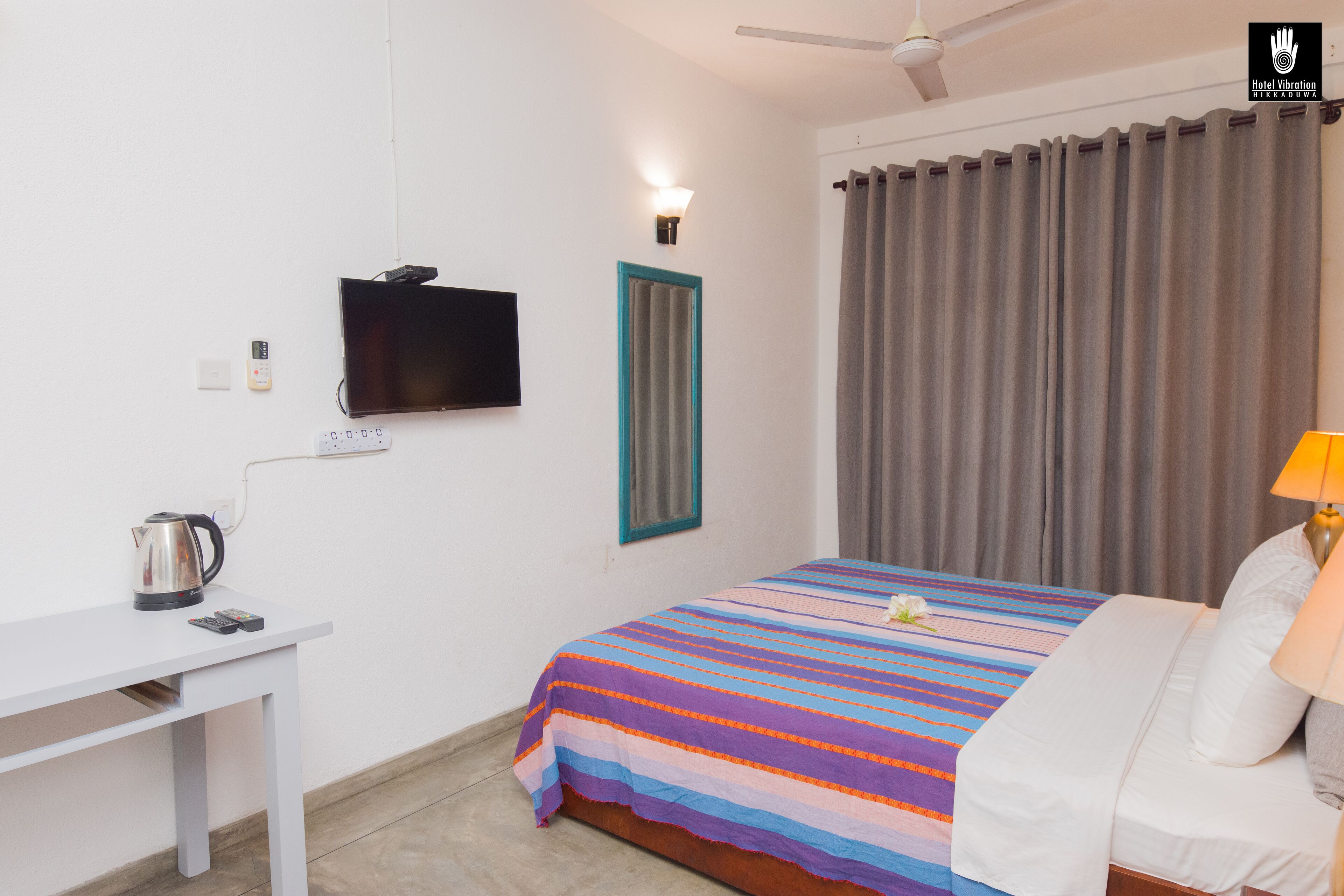 A hotel room with a bed, a desk with a kettle, a television, and a window with curtains.