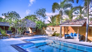 Piscina con sillones de descanso y un bote, rodeada de palmeras y edificios con tejados de tejas.