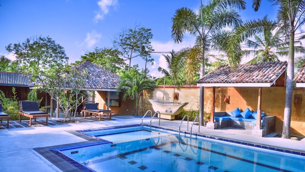 A swimming pool with lounge chairs and a boat, surrounded by palm trees and buildings with tiled roofs. - Hotel Vibration (Hikkaduwa)