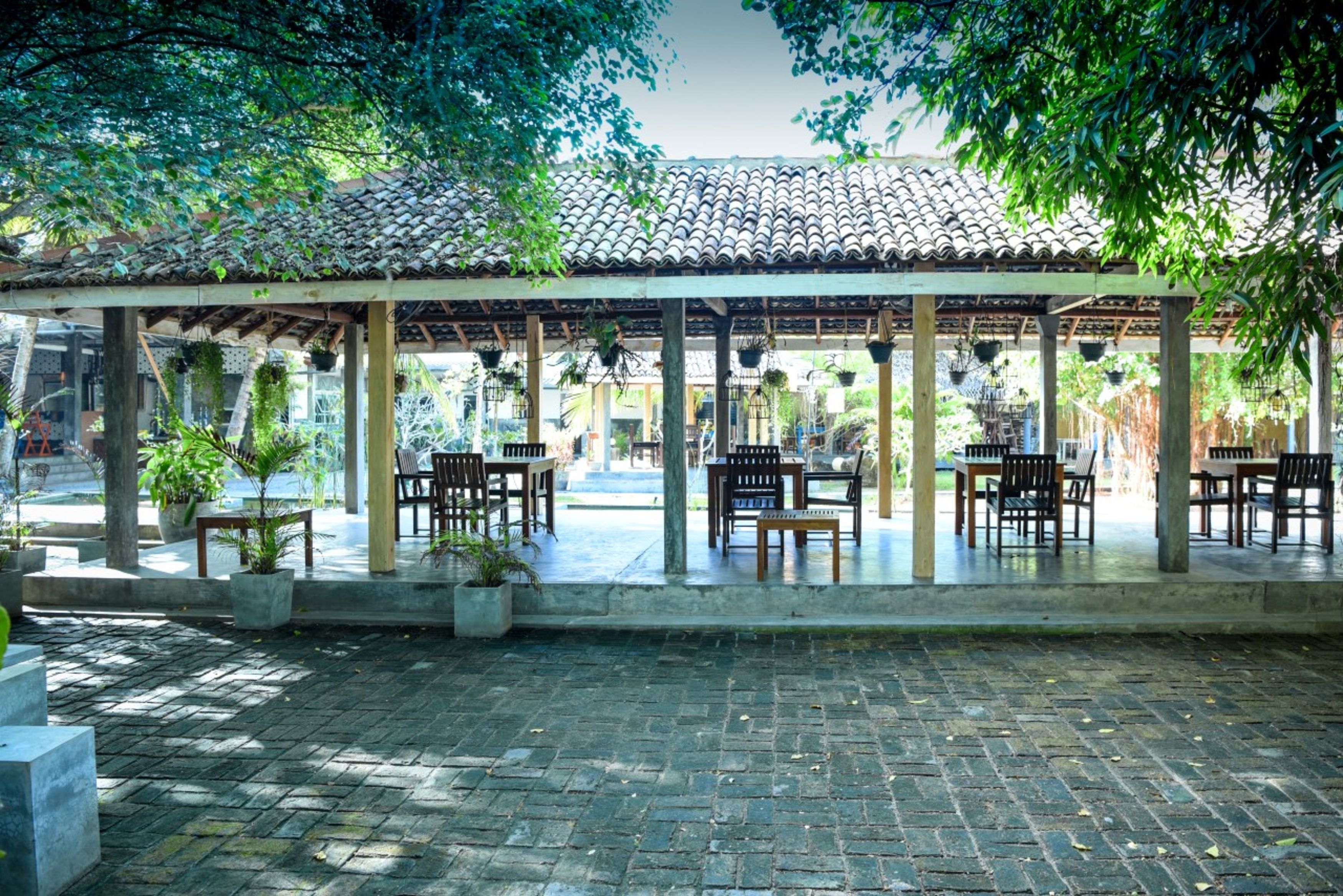 An outdoor covered patio area with brick flooring, wooden tables and chairs, and hanging plants.