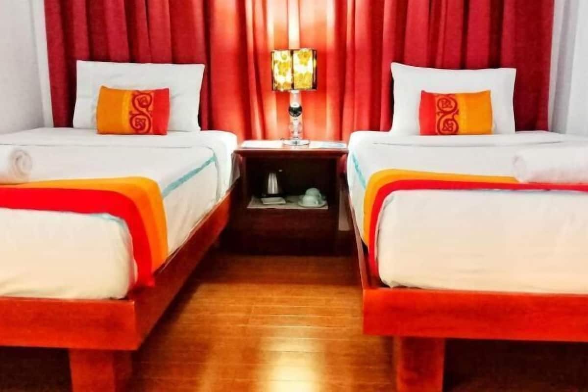 Quarto de hotel com duas camas, uma mesa de cabeceira com abajur e piso de madeira.