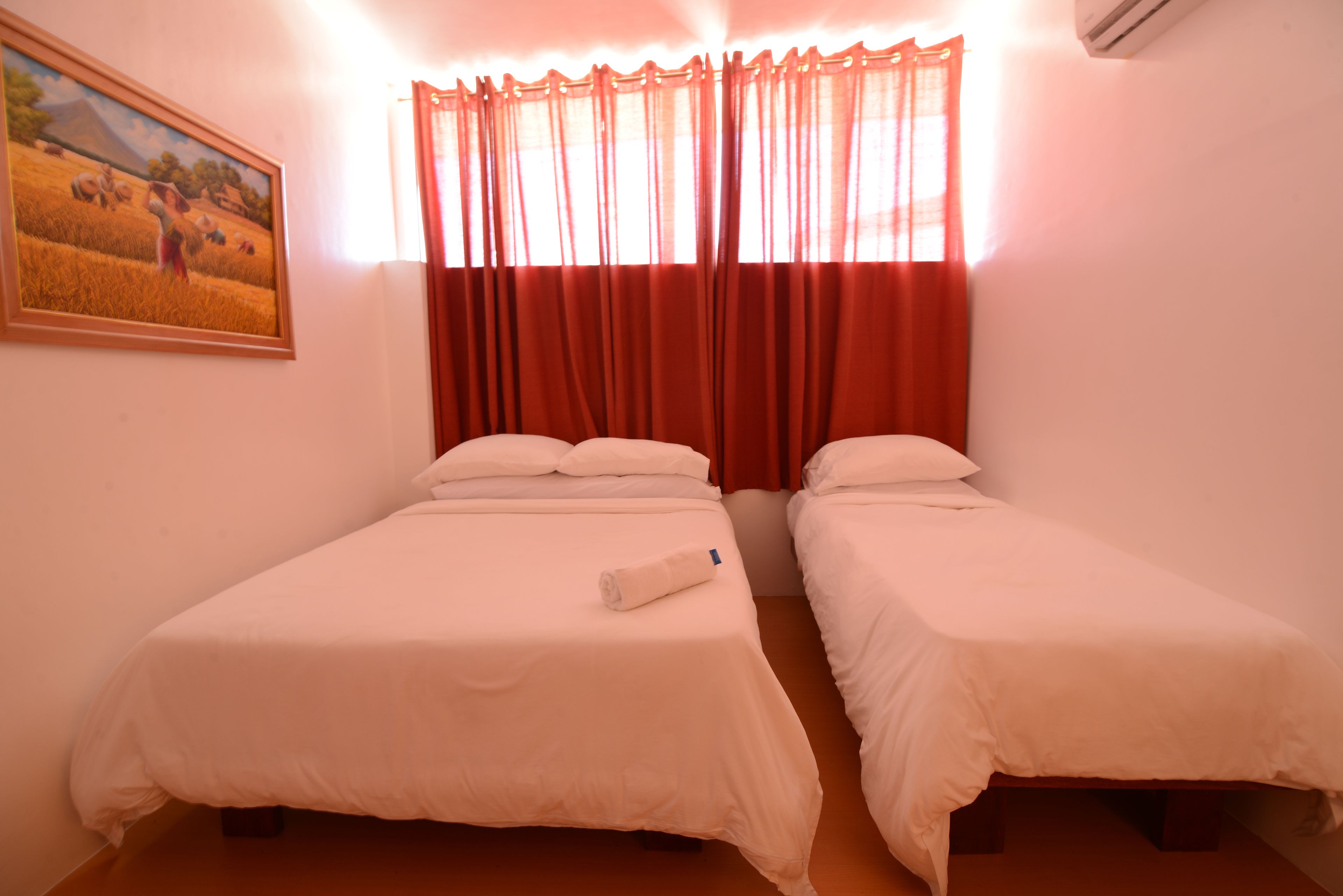 Une chambre avec deux lits simples, un tableau au mur et une fenêtre avec des rideaux rouges.