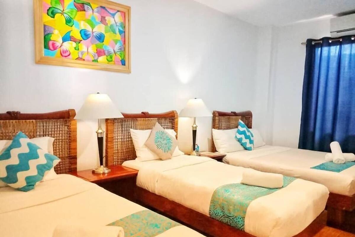 Quarto de hotel com três camas, cada uma com cabeceira de madeira, roupa de cama branca e almofadas decorativas. Há uma pintura abstrata colorida na parede acima das camas.
