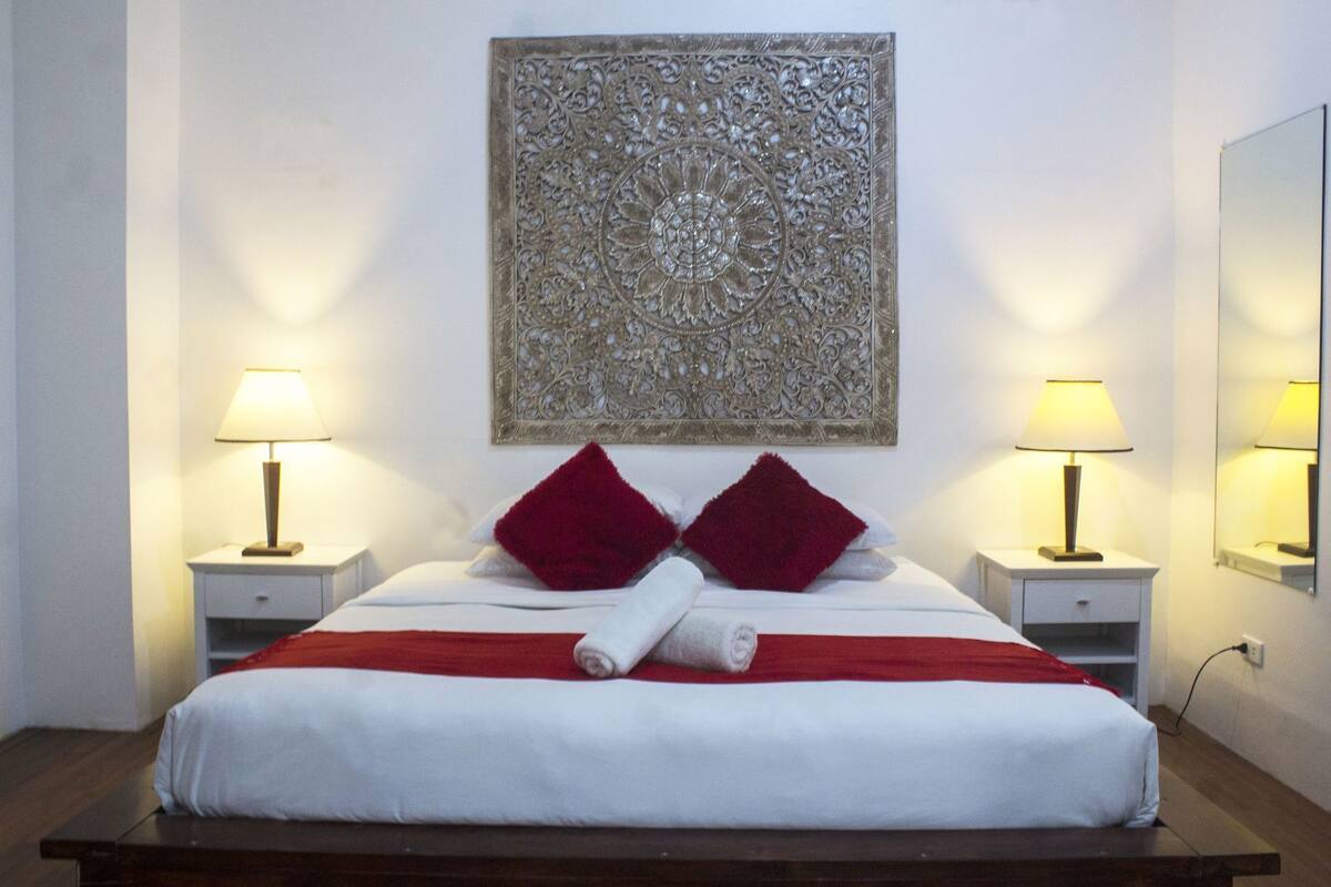 Uma cama bem arrumada com almofadas vermelhas, duas luminárias laterais e um adorno decorativo acima da cama.