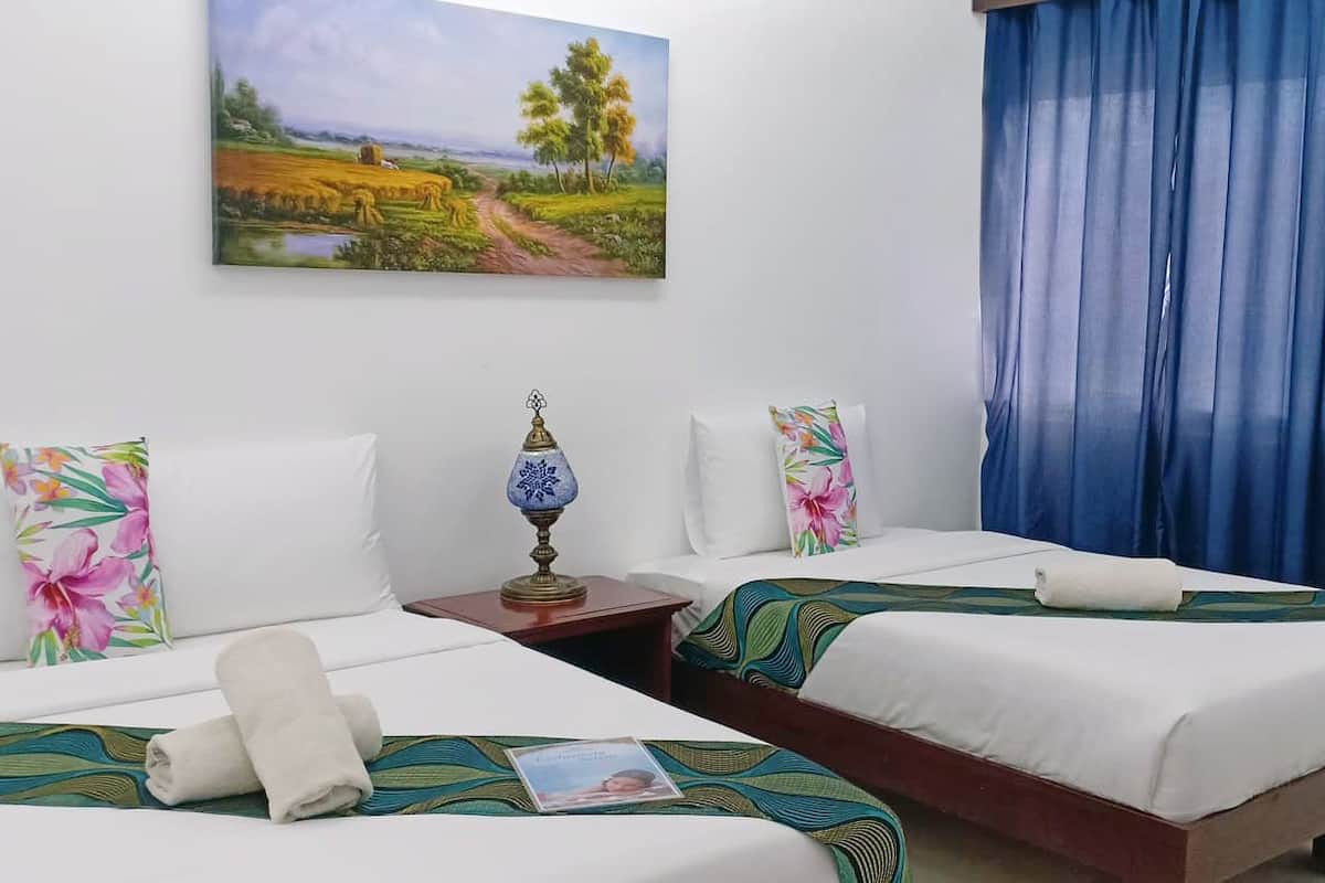 Quarto de hotel com duas camas, cada uma com colcha floral e travesseiro branco. Há um quadro na parede retratando uma paisagem, e uma cortina azul na janela.