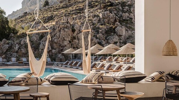 A poolside lounge area with seating and tables, a hanging chair, and a view of a rocky landscape.