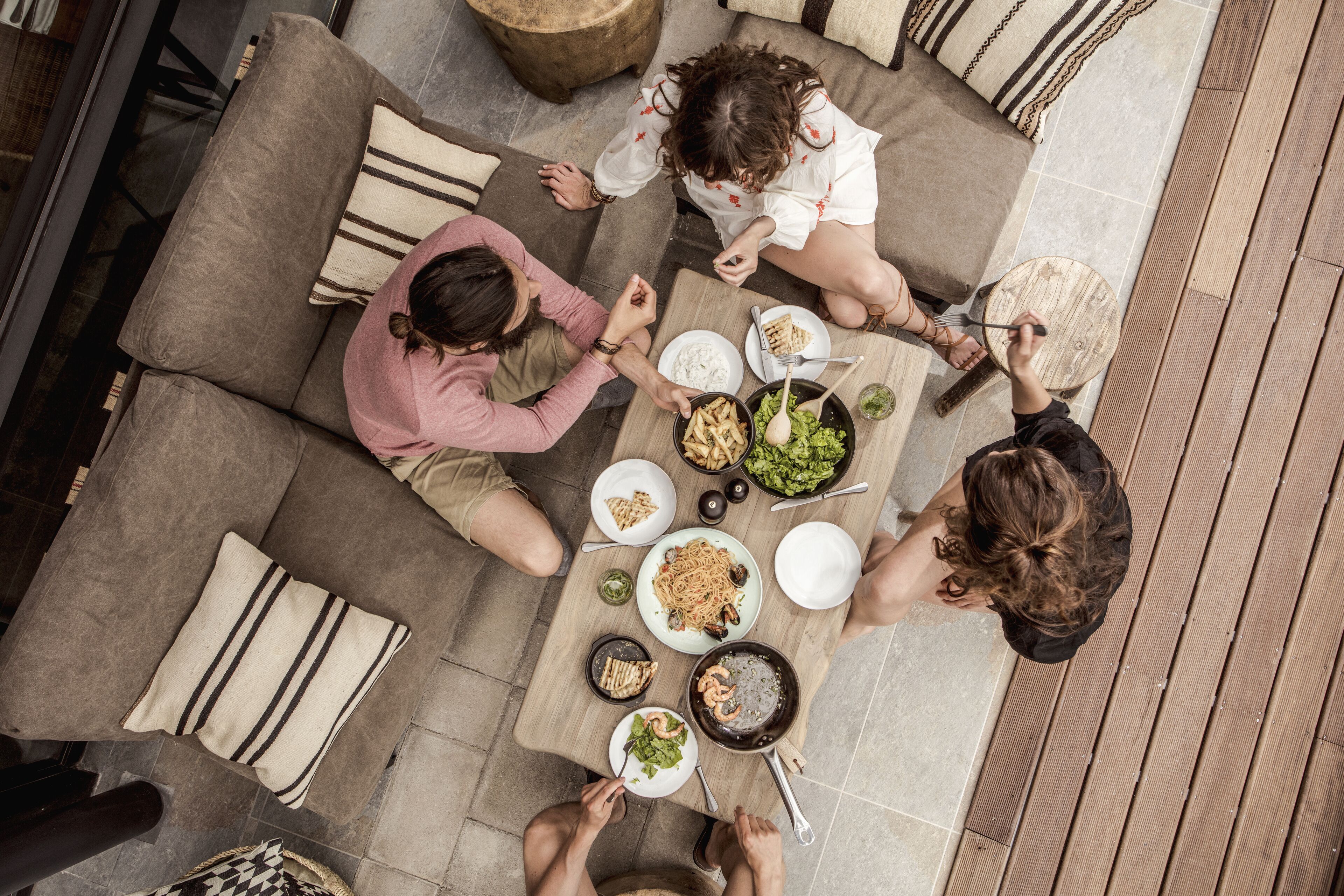 A group of people dining at an outdoor table with various dishes, surrounded by a wooden deck and a grey sofa.