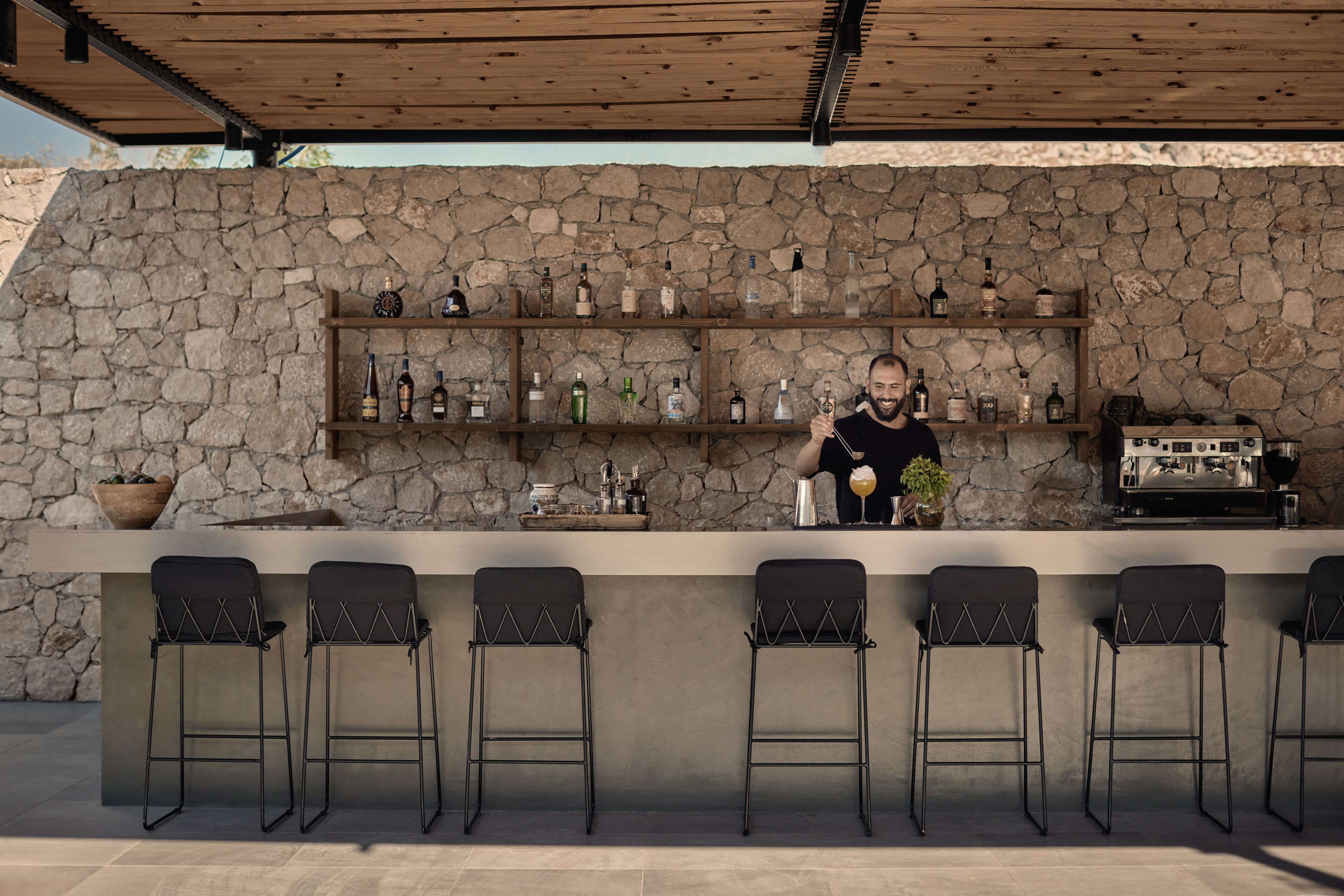 Een openluchtbar met een achtergrond van een stenen muur, meerdere flessenplanken en barstoelen.