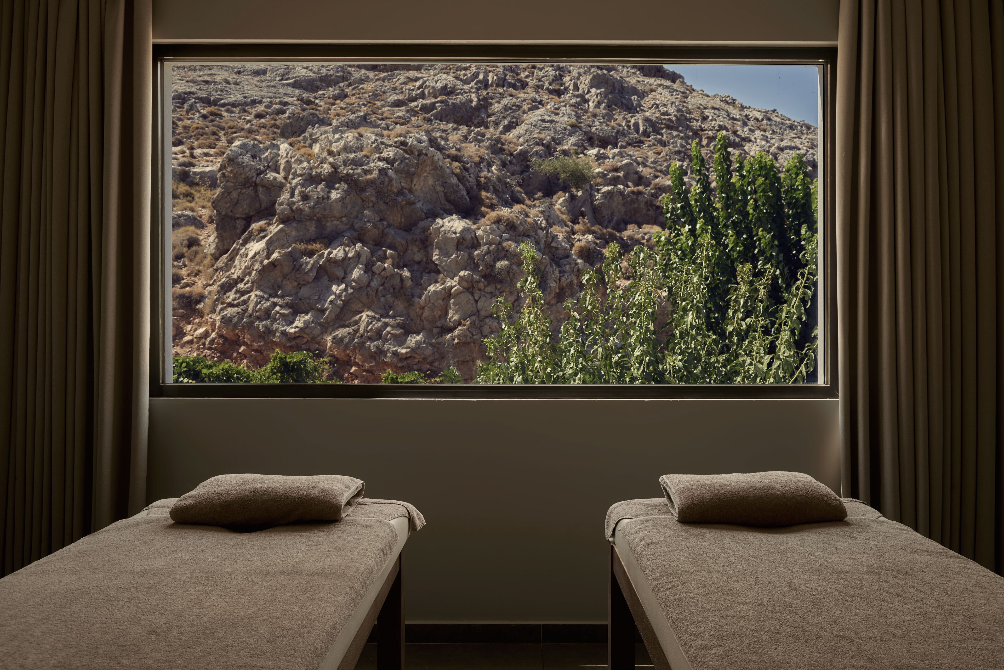 A room with two beds, a large window with a rocky landscape view, and curtains.