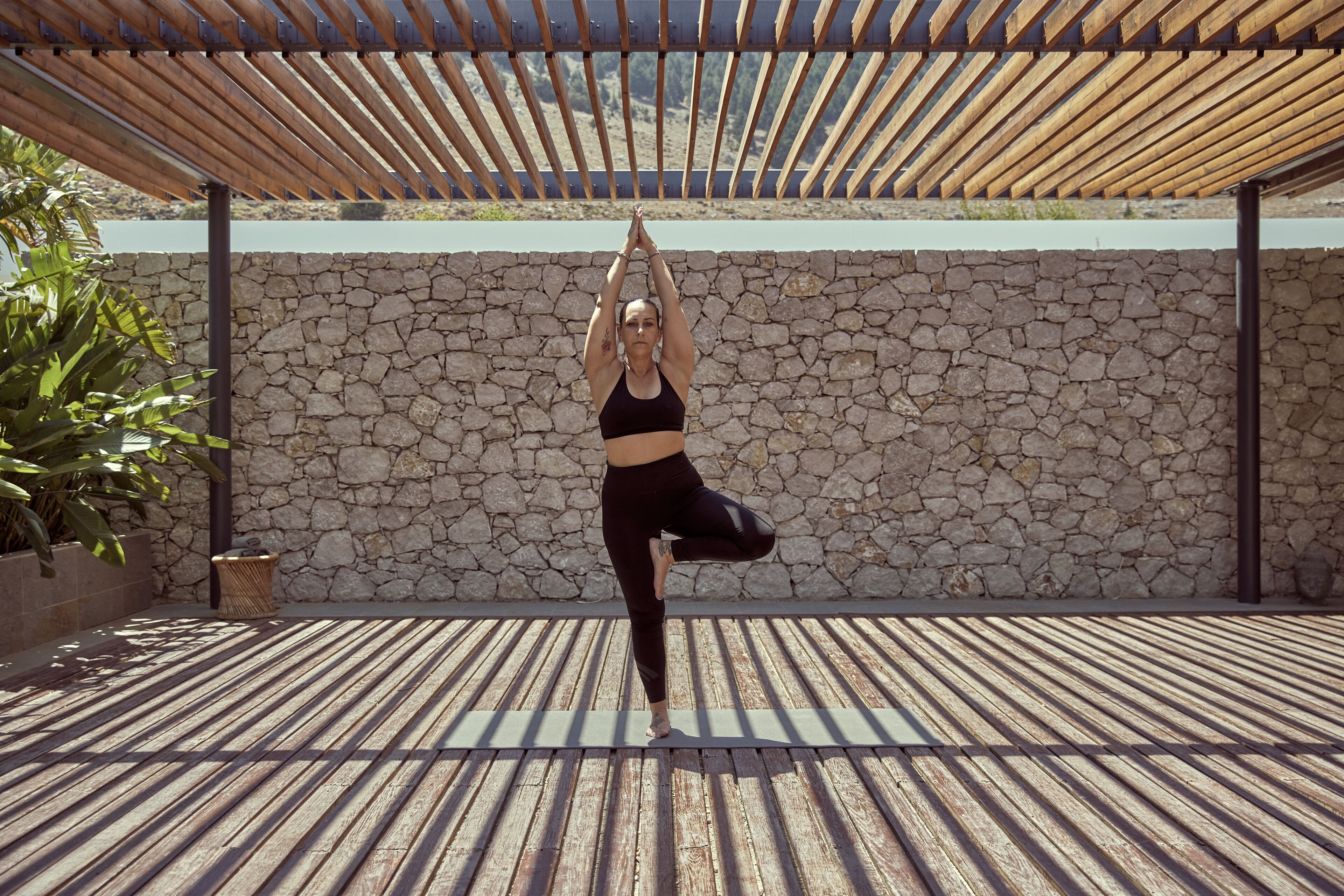 A person performing a yoga pose on a wooden deck with a stone wall and a pergola overhead.