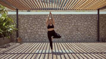 A person performing a yoga pose on a wooden deck with a stone wall and a pergola overhead.