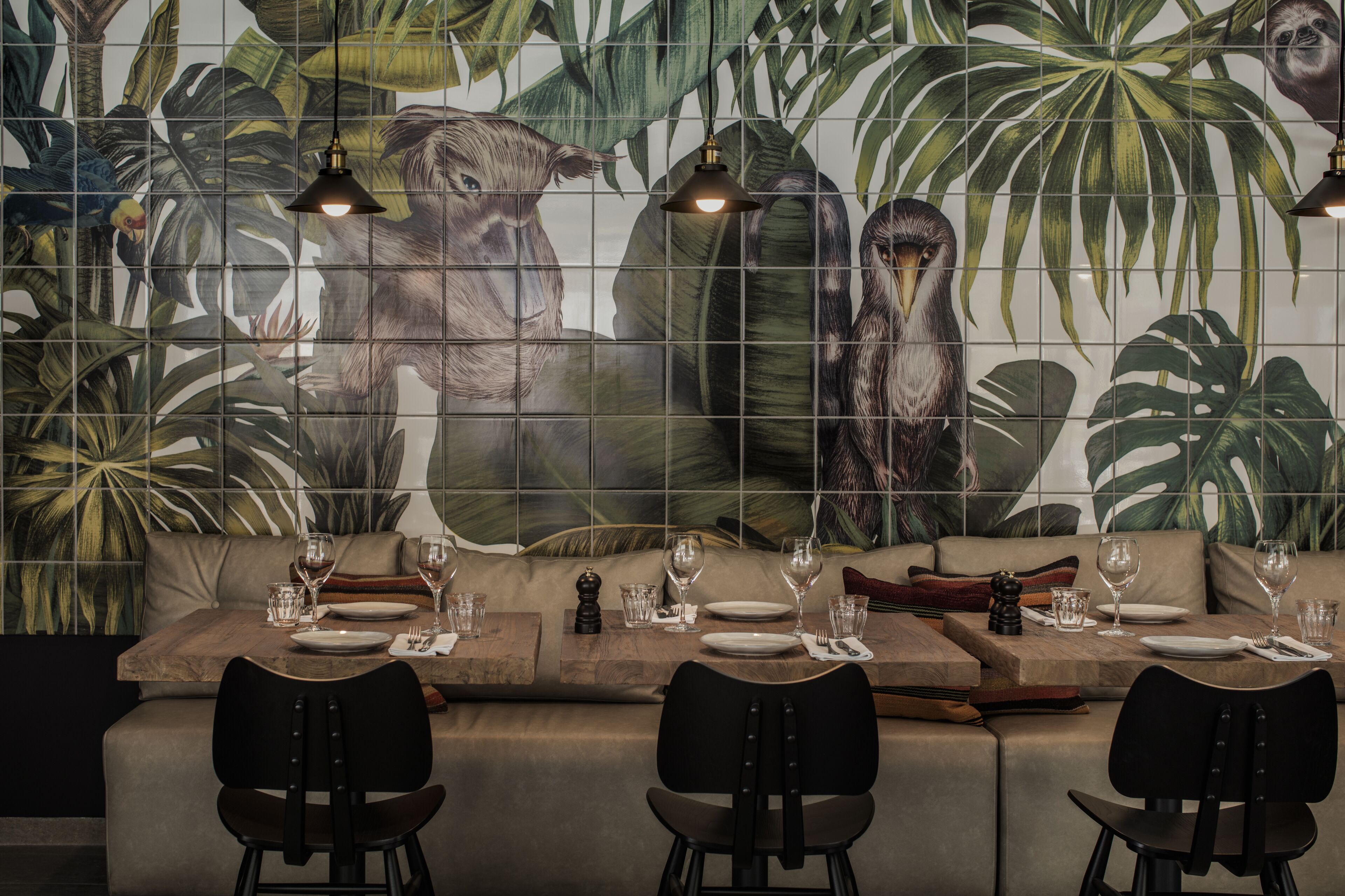 A restaurant with a mural of tropical foliage and animals on the wall, a long wooden table set for dining, and black chairs.