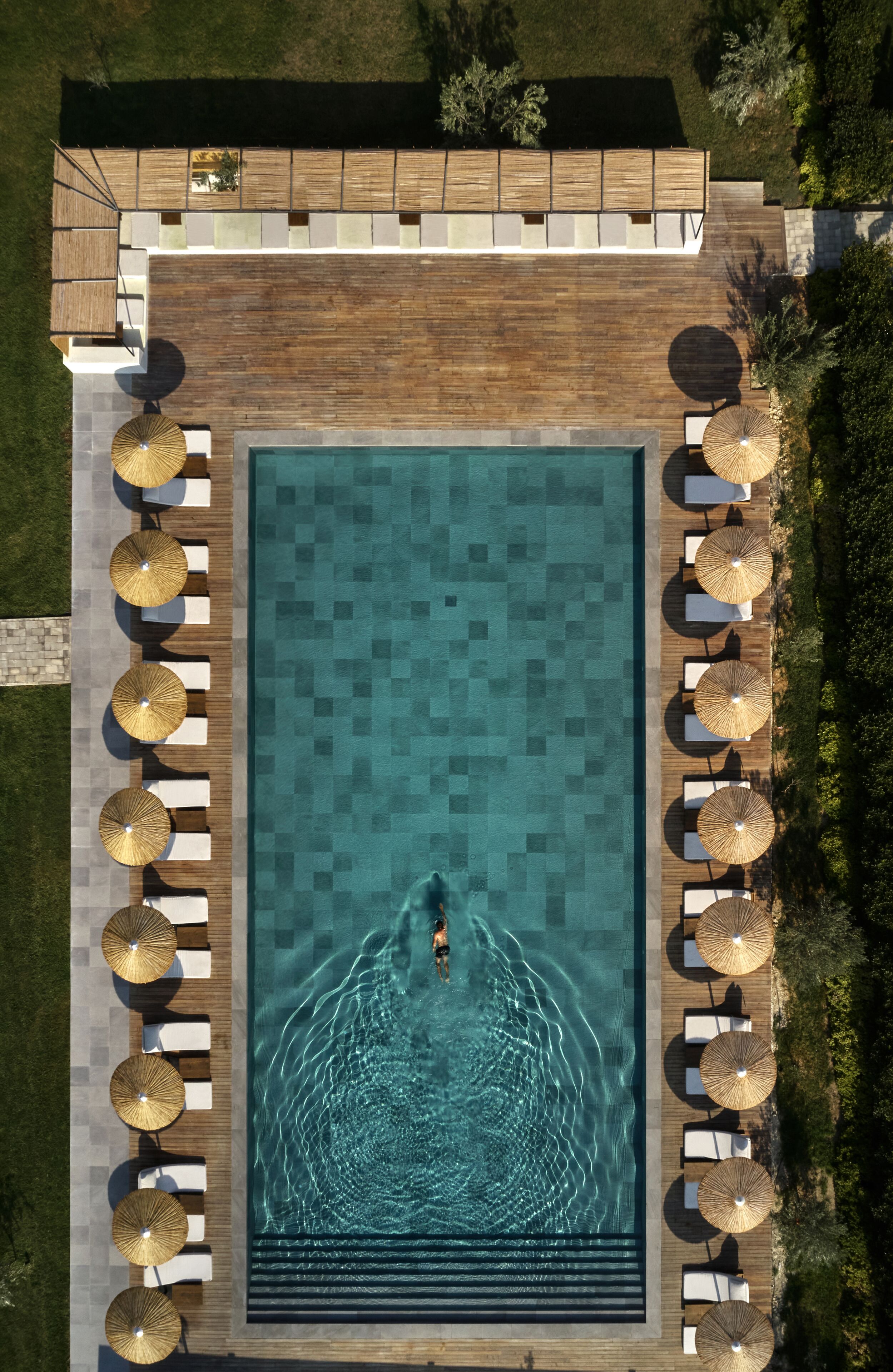 Vue aérienne d’une piscine rectangulaire entourée d’une terrasse en bois, agrémentée de chaises longues et de parasols.