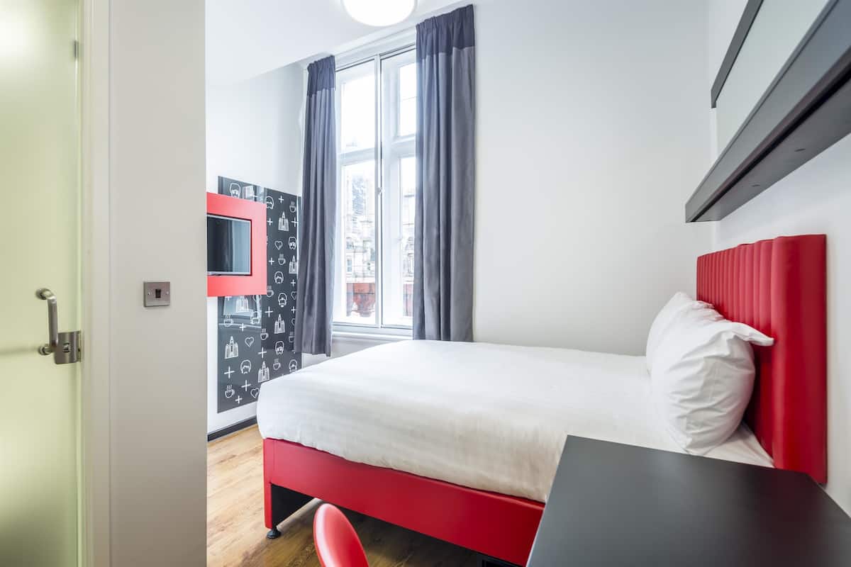 A modern hotel room with a bed, a desk, a window with curtains, and a wall with icons.