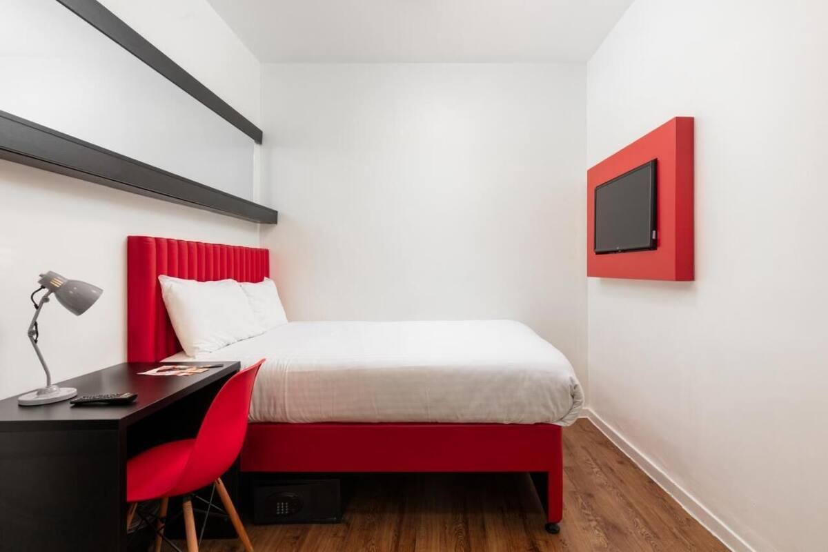 A small, modern bedroom with a red bed, white walls, and a black desk with a red chair.