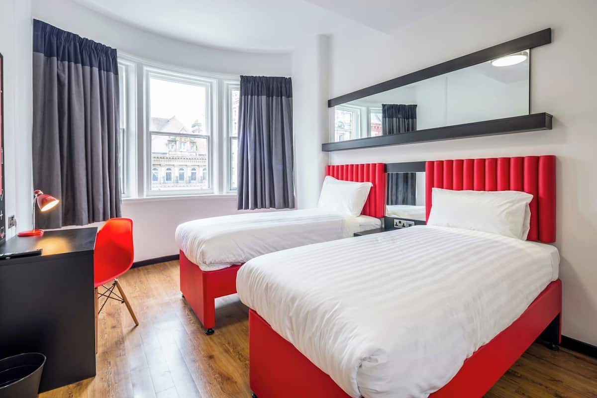 A hotel room with two beds, a desk, a chair, a desk lamp, and a window with curtains.