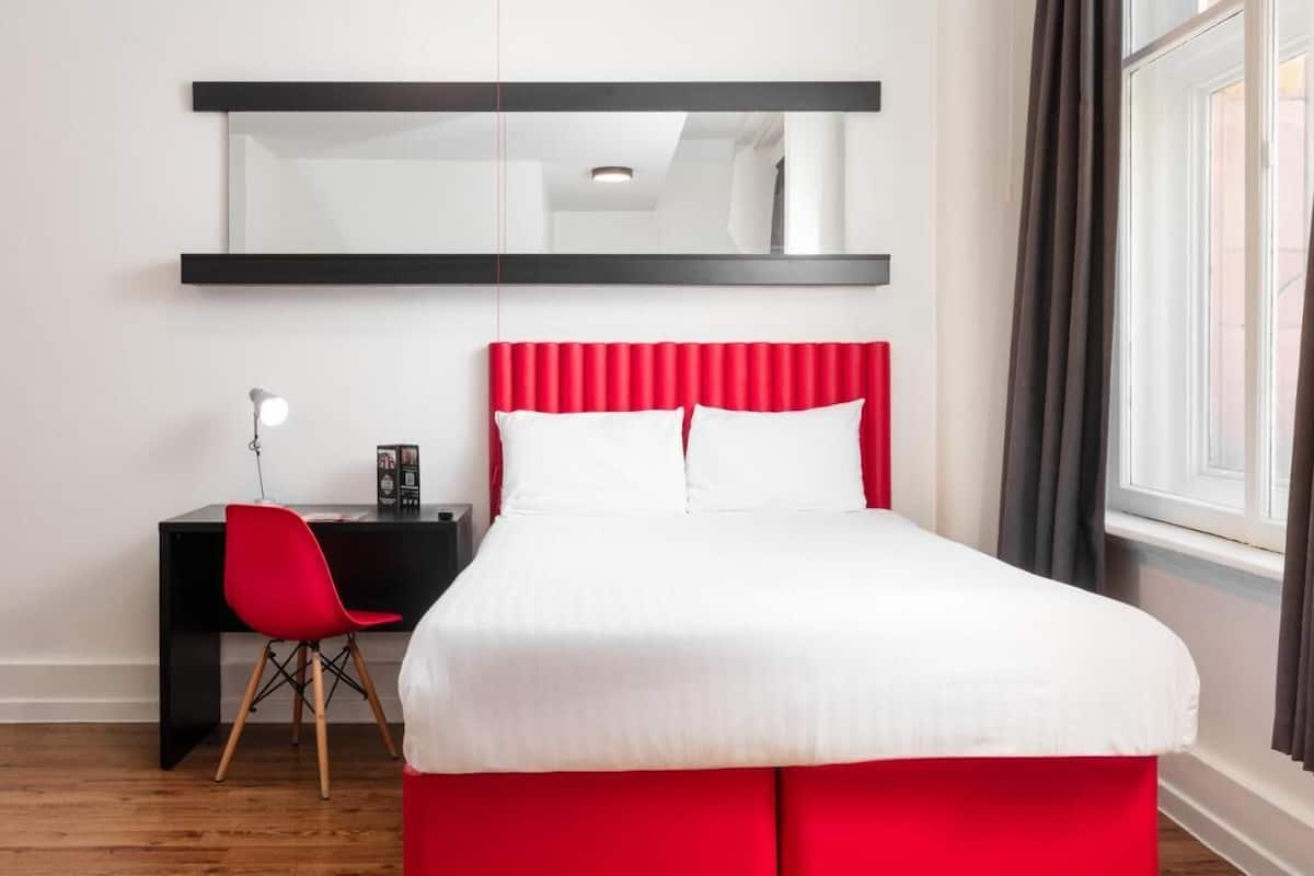 A modern hotel room with a red upholstered bed, a black desk with a red chair, a large mirror above the bed, and a window with curtains.