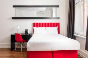 A modern hotel room with a red upholstered bed, a black desk with a red chair, a large mirror above the bed, and a window with curtains.