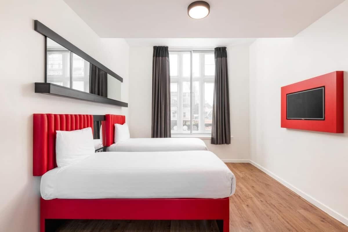 A hotel room with two beds, a large window with curtains, a mounted TV, and a modern light fixture.