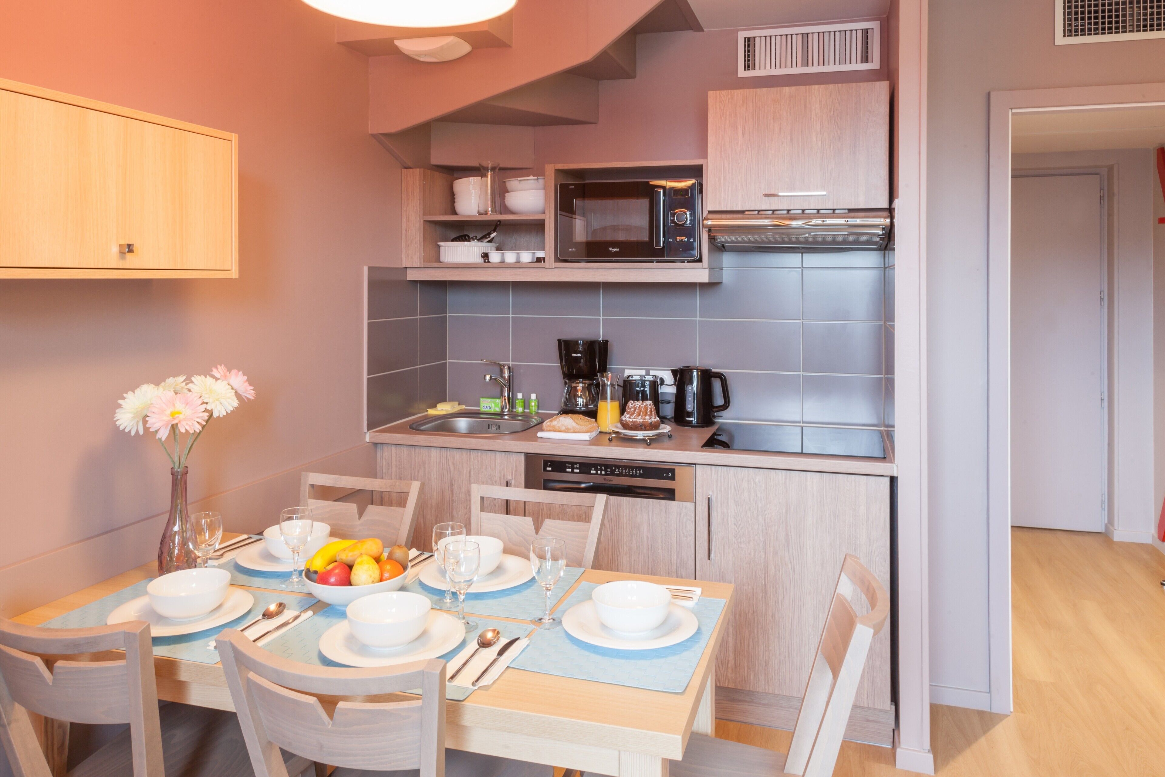 A compact kitchen with a microwave, coffee maker, and dishwasher. There is a dining table set with plates, bowls, and cutlery.