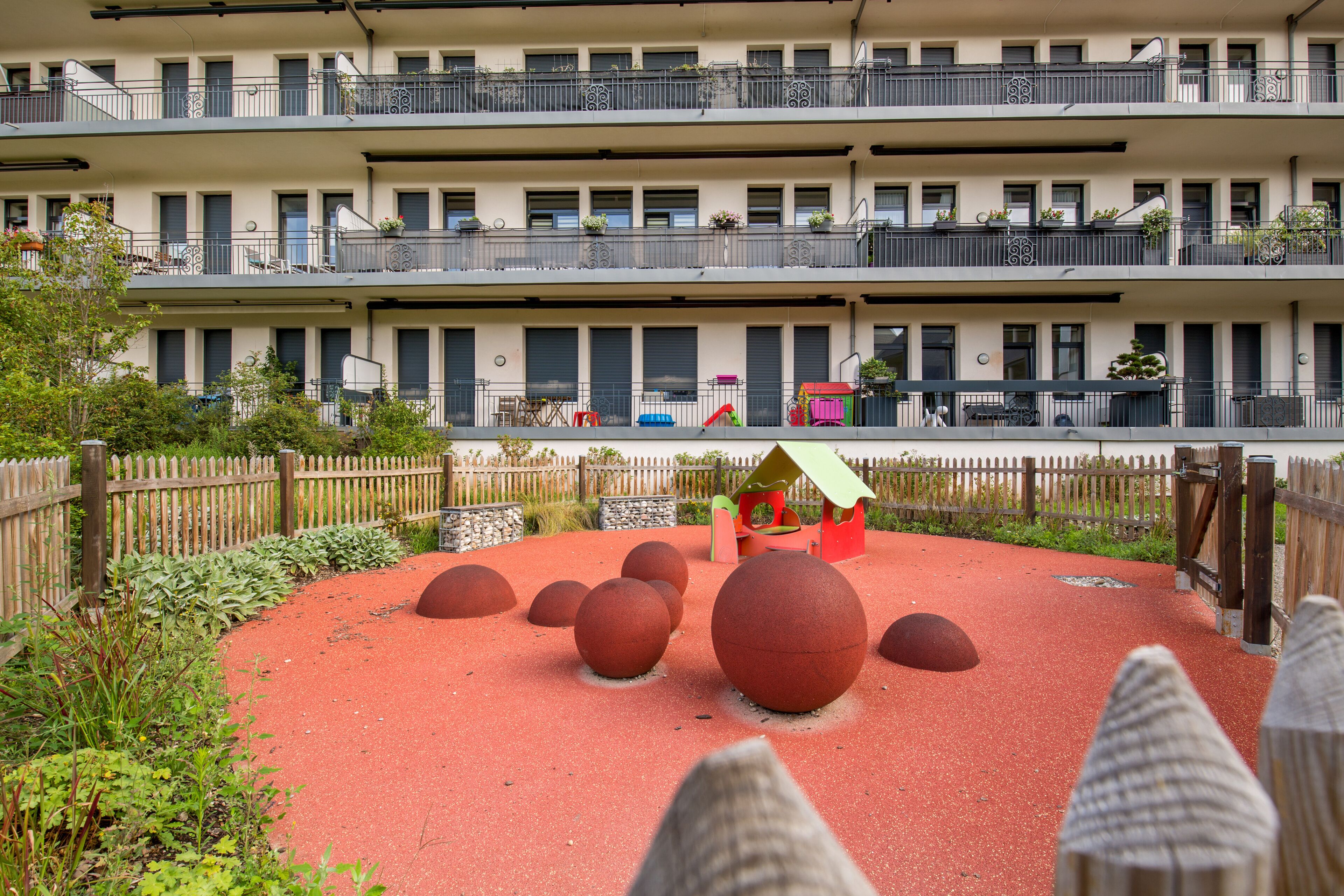 A multi-story building with balconies, a playground with red balls, and a wooden fence.