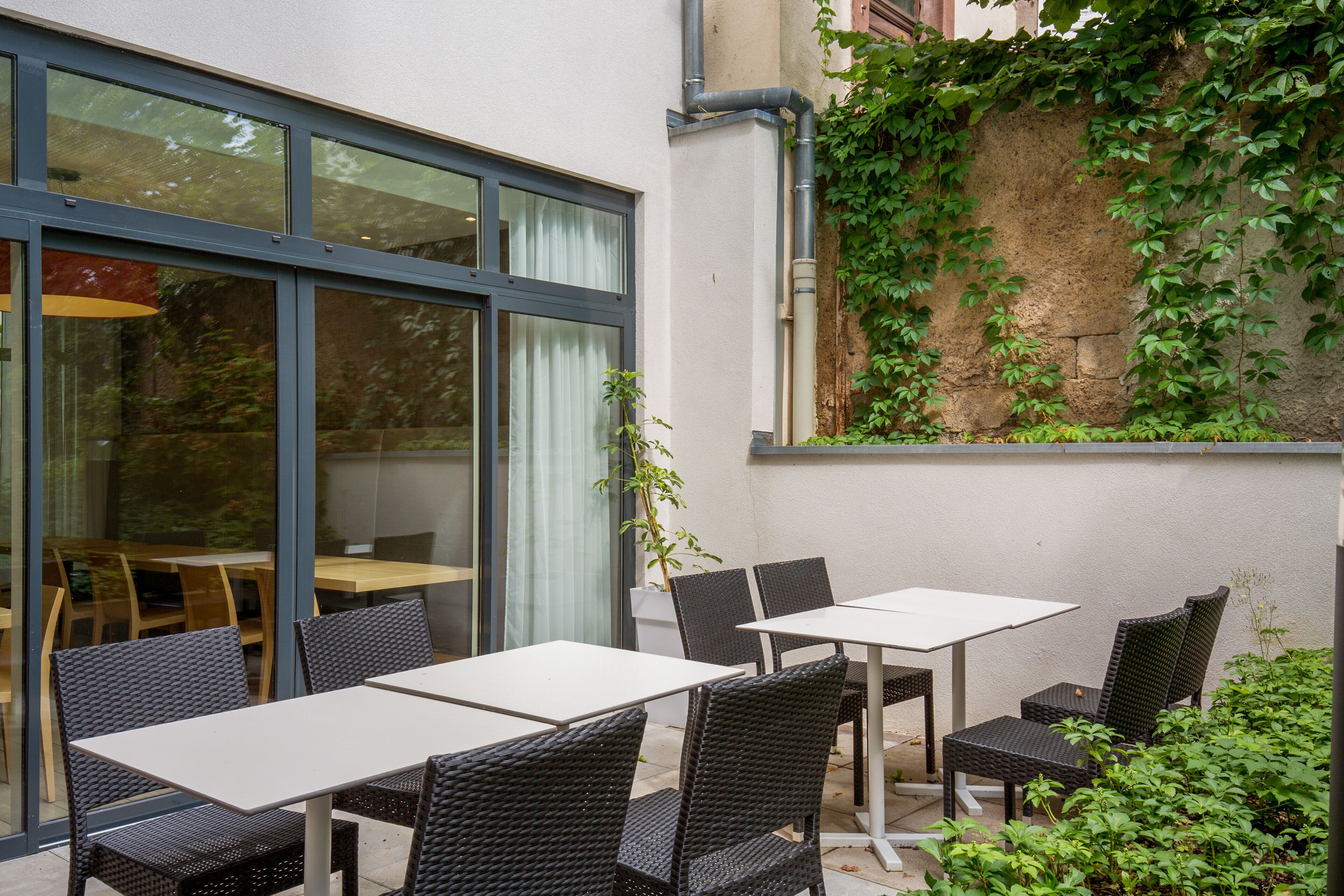 Une terrasse extérieure avec des tables et des chaises, une entrée avec une porte vitrée et un mur recouvert de plantes grimpantes.