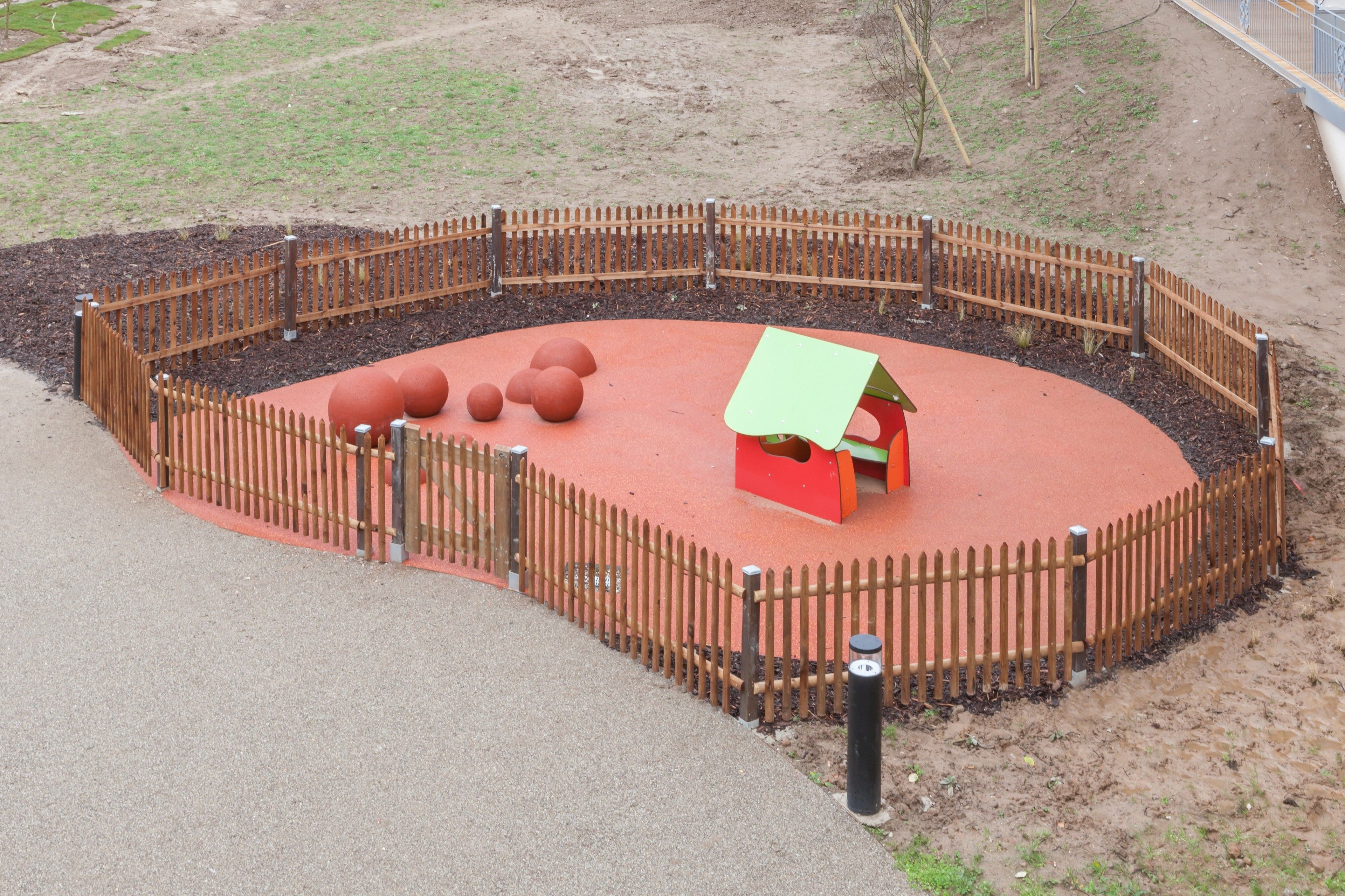 A fenced outdoor play area with a red surface, a small red house, and several large orange balls.
