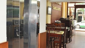 A hotel lobby with a reception desk, wooden chairs, a glass elevator, and a tiled floor.