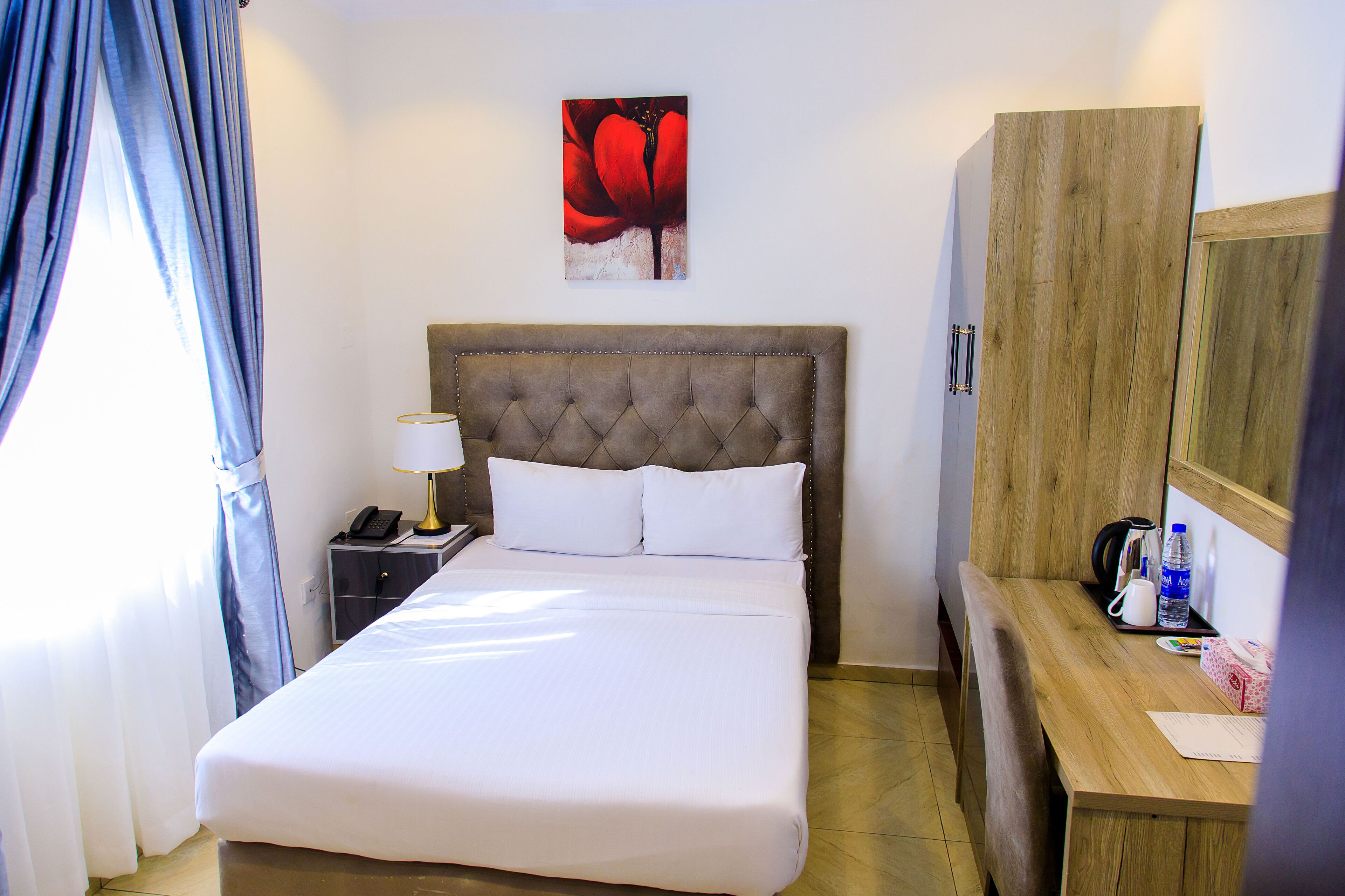 A hotel room with a large bed, a bedside table with a lamp, a wooden wardrobe, and a painting on the wall.