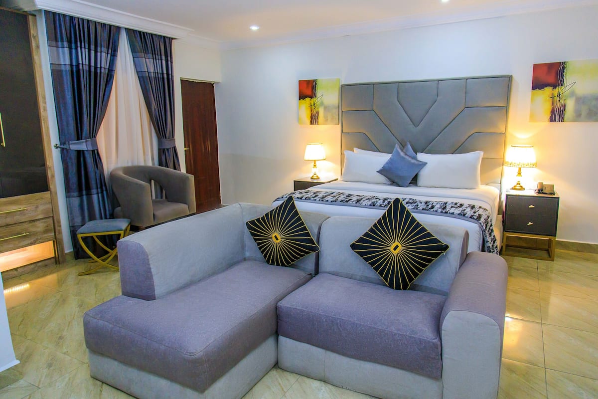 A modern hotel room with a large bed, two bedside tables with lamps, a grey armchair, and a sofa with decorative pillows.