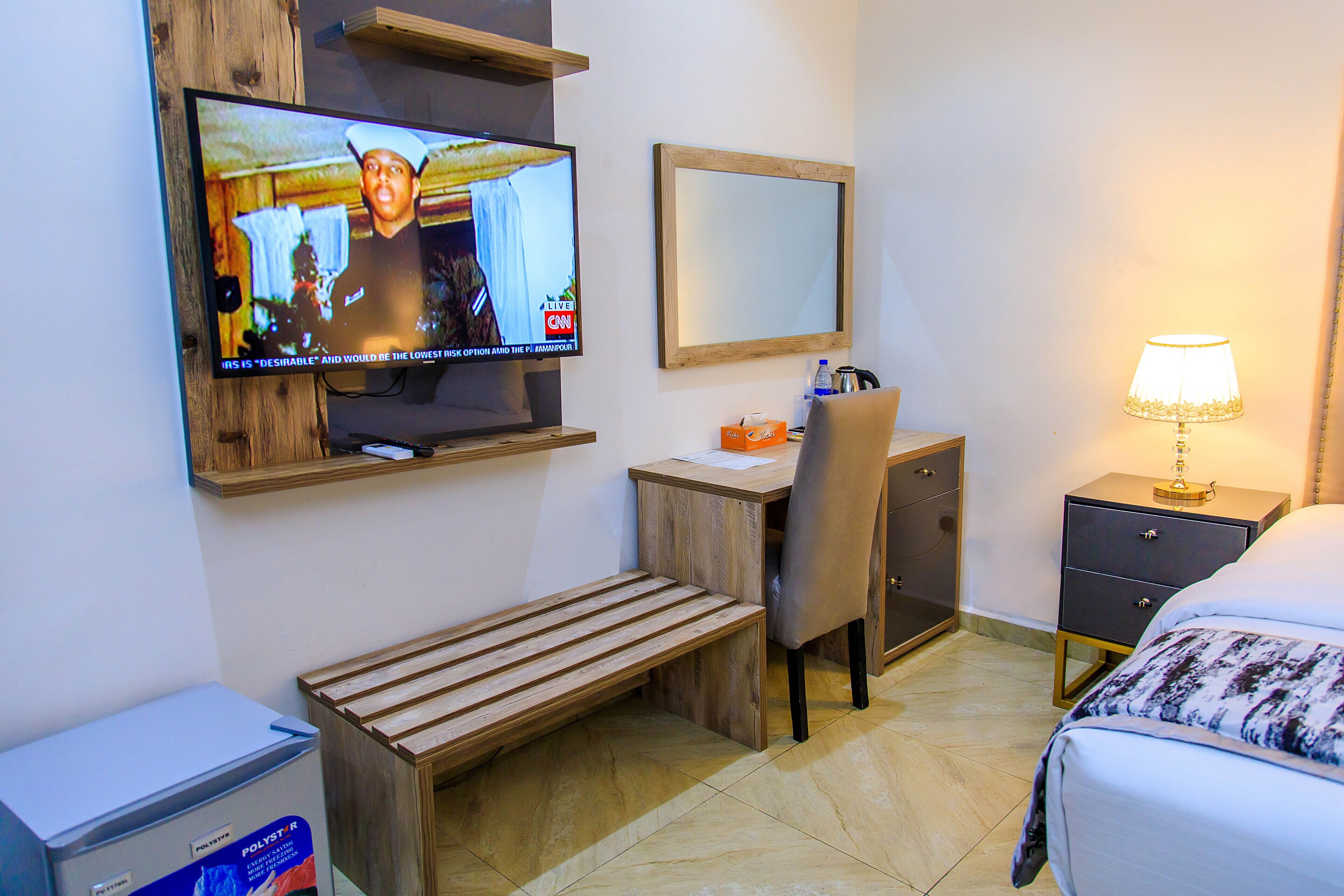 Camera d'albergo con televisione a parete, una scrivania con sedia, comodino con lampada, un letto e una panca in legno.