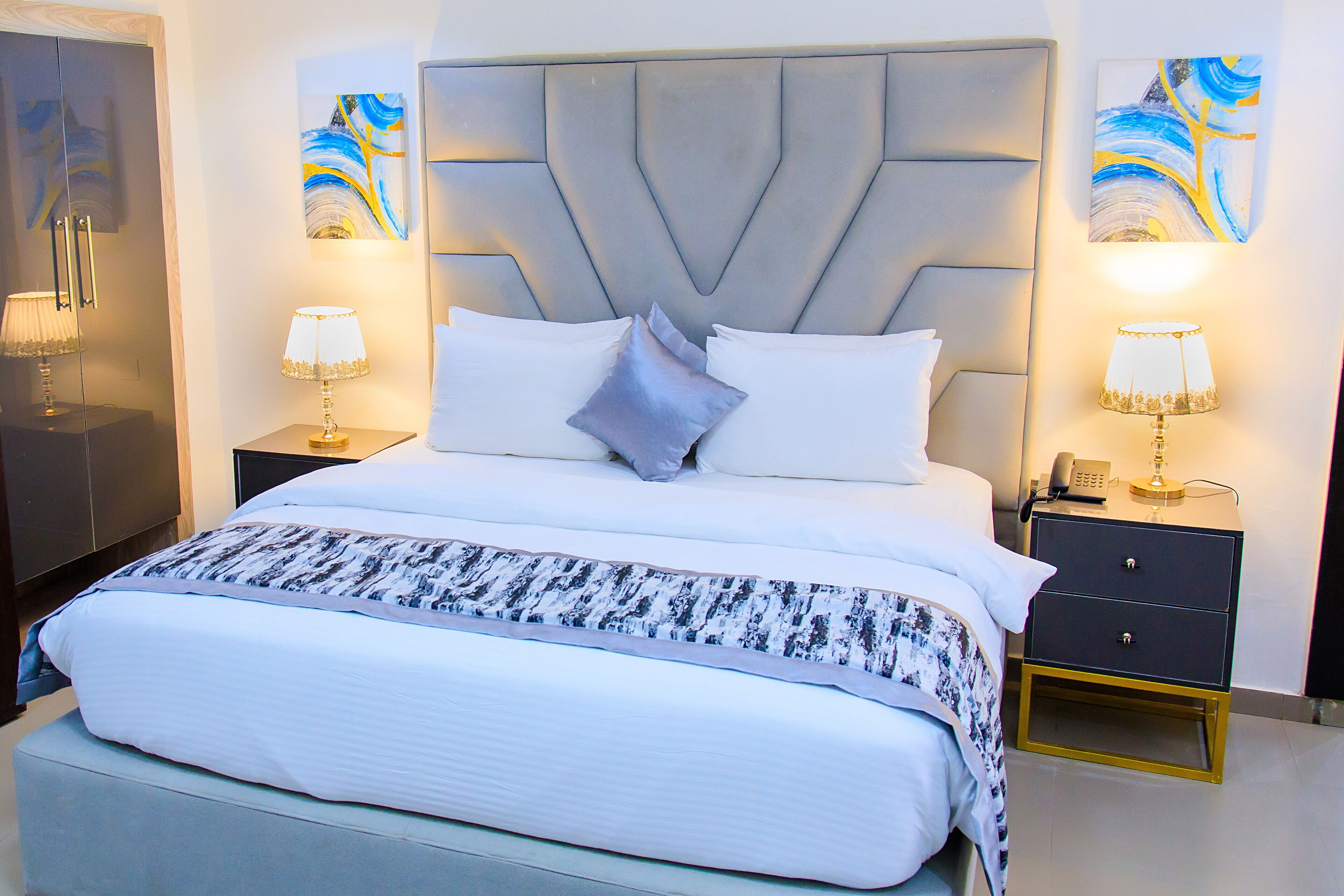 A modern hotel room with a large bed, two bedside tables, a wardrobe, and abstract wall art.