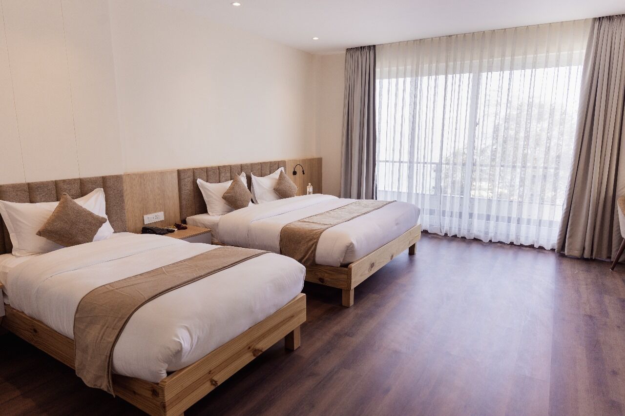 Quarto de hotel com duas camas de madeira, uma janela grande com cortinas finas e piso de madeira.