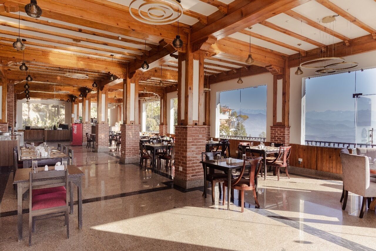 A spacious restaurant with wooden beams and brick pillars, large windows offering mountain views, and a variety of seating arrangements.